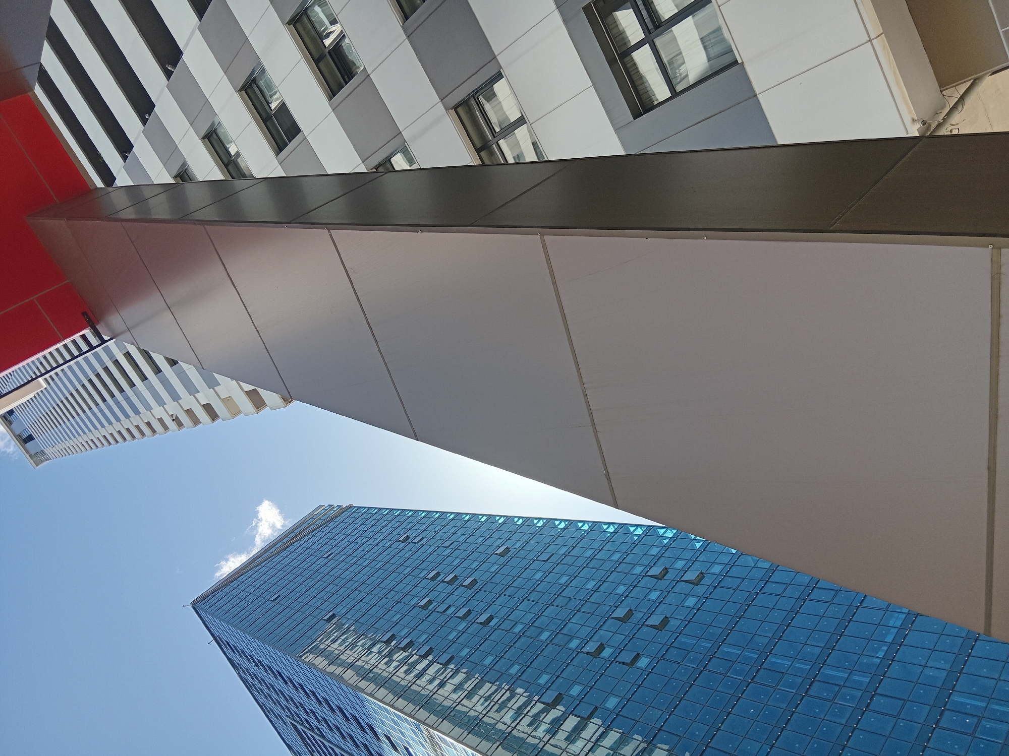 The image shows a low-angle view of several modern buildings in Batumi, Georgia. A pedestrian bridge is visible between two of the buildings. The building on the lower right appears to be a skyscraper with a blue glass facade. The sky is bright blue with a few clouds. The buildings and bridge create a sharp contrast in shapes and colors, with the blue skyscraper, the gray bridge, and other buildings with red and white details.