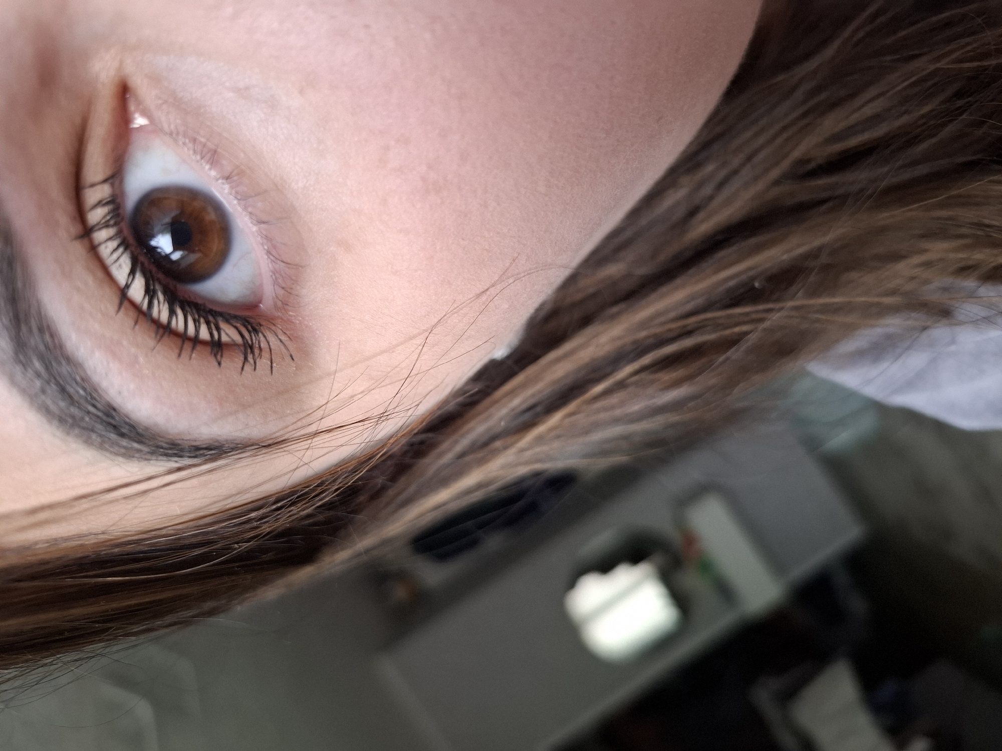 A close-up, tilted image features a woman's brown eye with dark eyelashes enhanced by mascara. Above the eye, a thick, dark eyebrow is visible. The skin is light-toned and smooth. Brown hair is partially visible, framing the eye and face. In the background, a blurry and undiscernible room interior can be vaguely seen. The location or city cannot be definitively determined from the image alone.