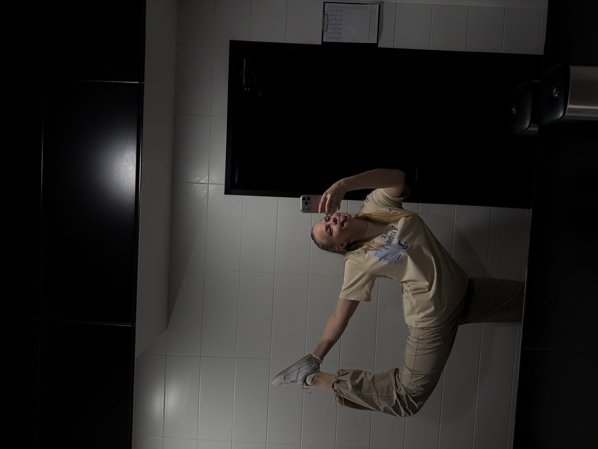 A young woman with blonde hair pulled back is performing a backbend in a restroom. She is wearing a light-colored t-shirt and loose-fitting beige pants. Her right foot is bent and touching the back of her head, while her left leg is extended horizontally behind her. She is holding a pink smartphone in her right hand, taking a selfie, and sticking her tongue out at the camera.
The restroom has white tiled walls and a black partition in the background. A large black mirror reflects the scene. On the right wall, a white clipboard with a schedule is attached. In the foreground, two metal and black trash receptacles are visible.
The image is set in a restroom within the city of Izhevsk, Russia.