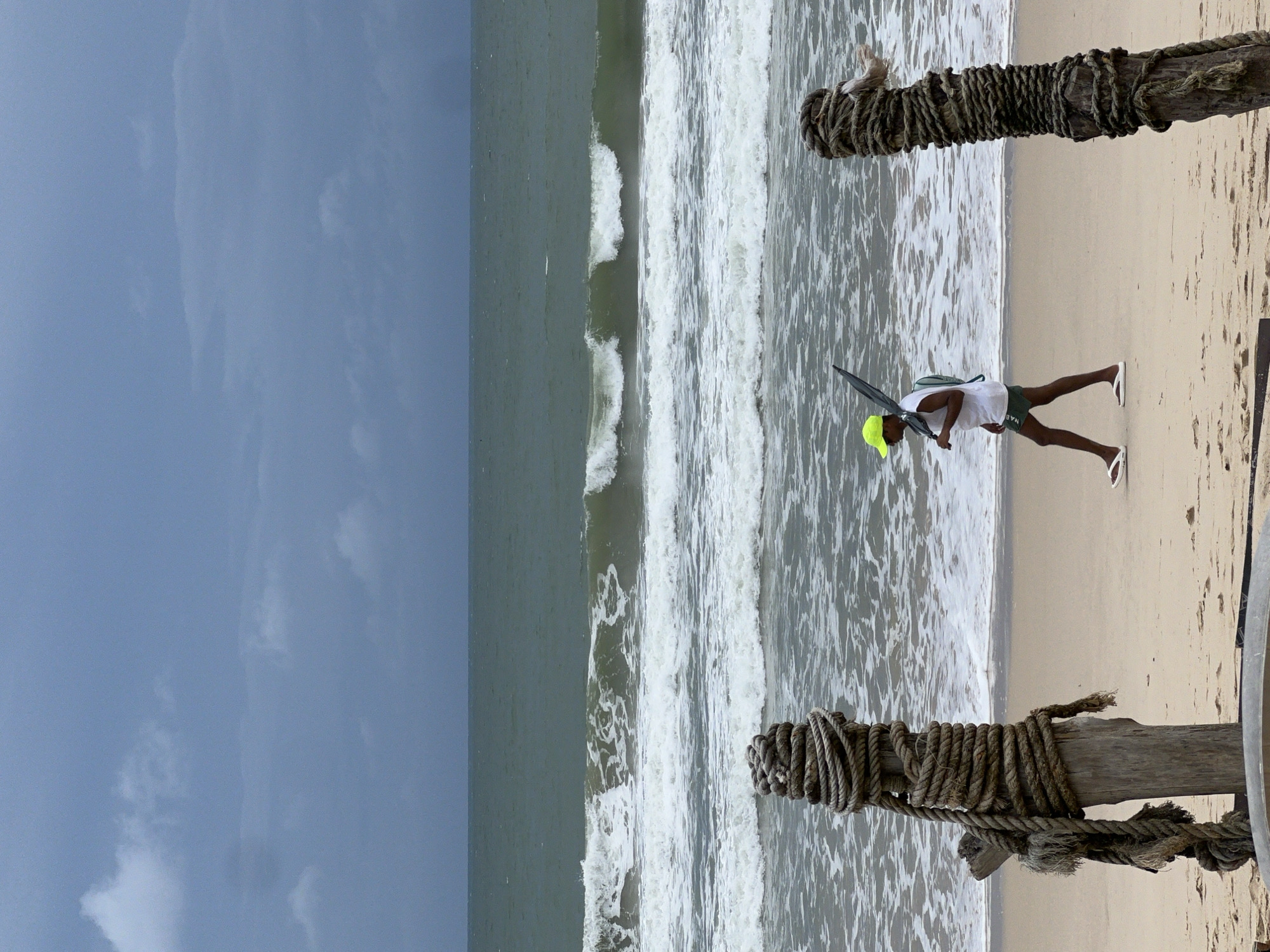 The image shows a beach scene on a possibly overcast day. A person is standing on the sand near the shoreline, holding a dark-colored umbrella or similar object. They are wearing a light-colored shirt, dark shorts, a bright yellow hat, and white flip-flops. The beach stretches from the lower right to the middle of the frame where waves are breaking. Two wooden posts wrapped in thick rope are positioned on the right side of the image, likely part of some structure, the beach is a medium beige color. The sky is overcast with grey clouds covering much of the left side of the image. The location appears to be near the city of Unknown, Unknown.