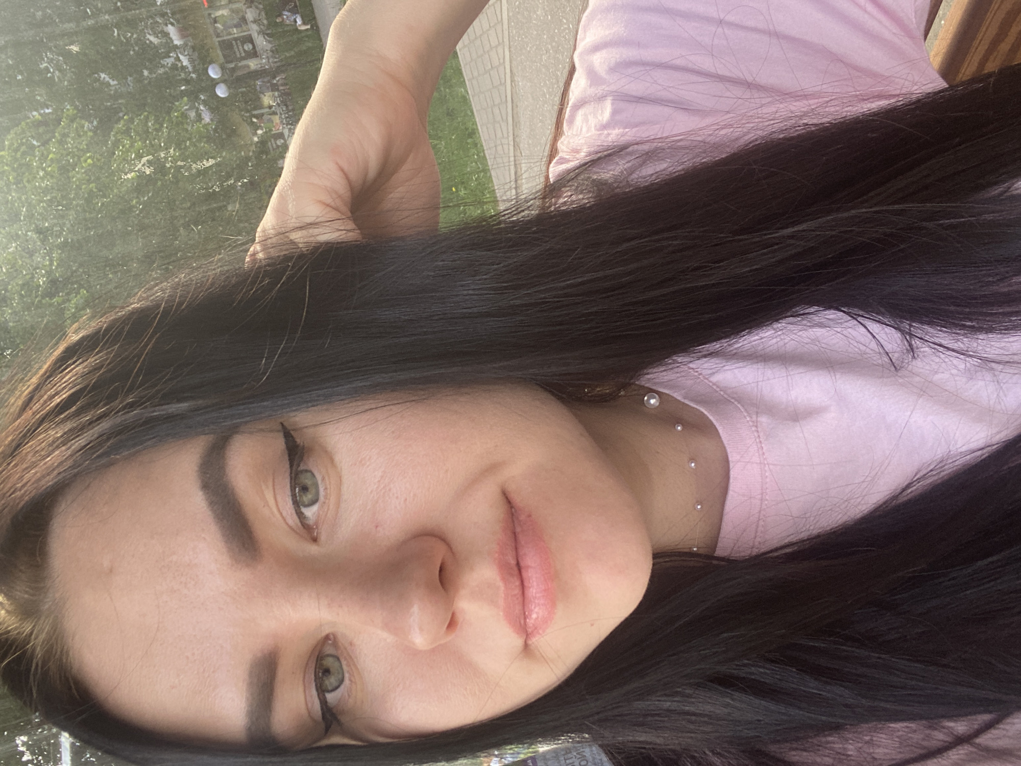 A woman is lying on a bench, facing the camera. She has long, dark hair, green eyes, and fair skin. She is wearing a light pink t-shirt and a pearl necklace. Her left hand is raised near her head. The background includes greenery, trees, and pavement. This image appears to be taken in a park or outdoor area in the city of Chaikovsky, Russia.