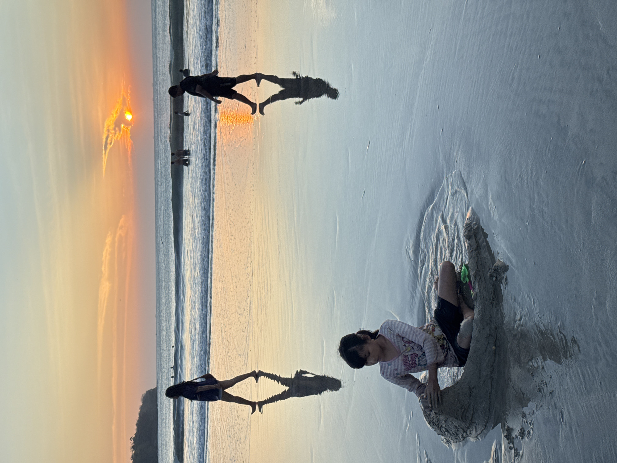 A young girl is building a sandcastle on the wet sand of Kuta Selatan beach in Bali, Indonesia, during sunset. She is sitting down, wearing a striped shirt and dark shorts, and is actively shaping the sand with her hands. In the background, the ocean stretches out with gentle waves, and the setting sun paints the sky in warm hues of orange and yellow. Silhouettes of people can be seen walking along the shoreline and swimming in the water. The wet sand reflects the colors of the sunset and the figures, creating a serene and picturesque scene.