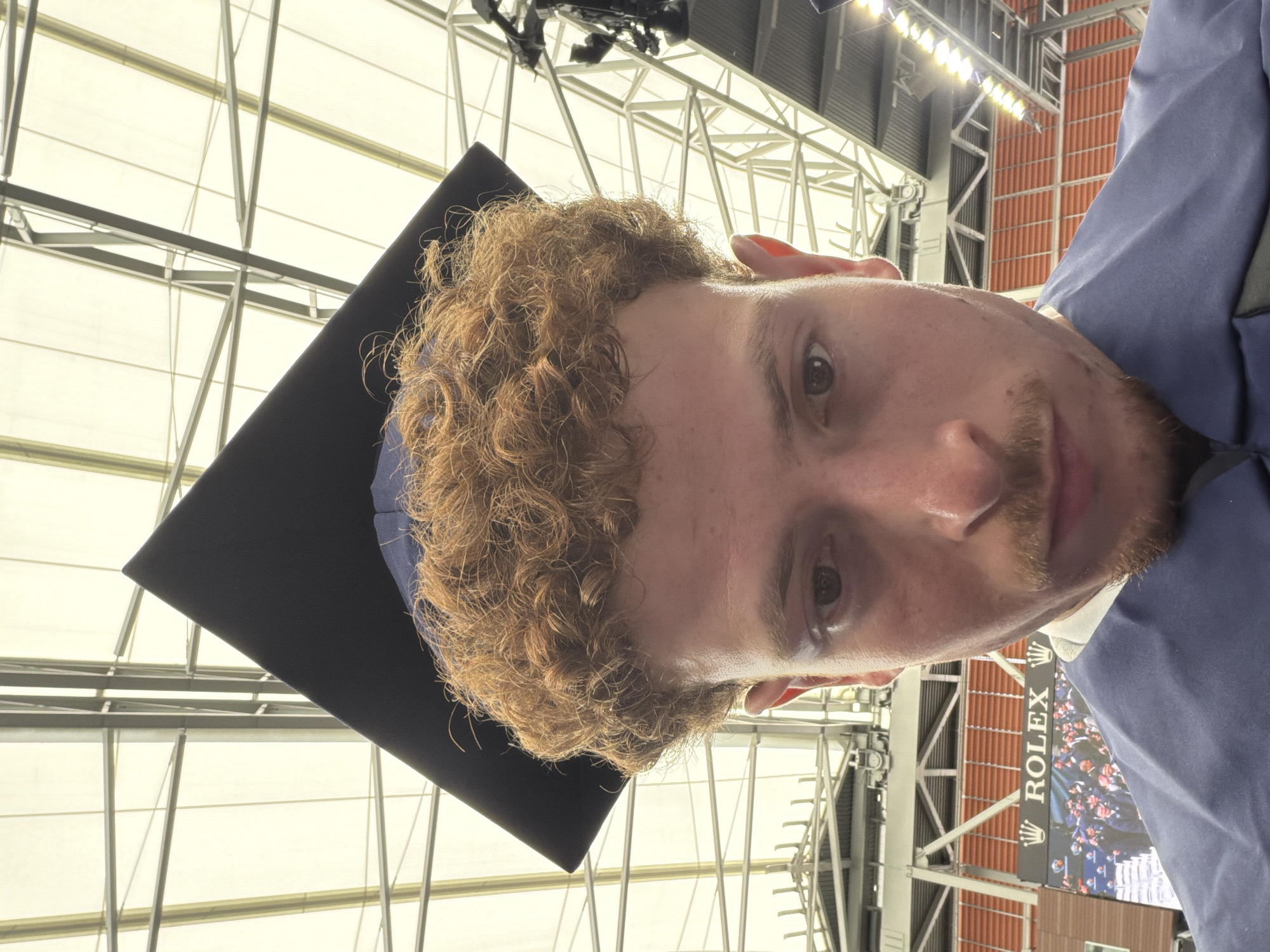 A man with curly, ginger hair is wearing a graduation cap and gown inside Arthur Ashe Stadium in New York City. The stadium's retractable roof and lighting are visible in the background. A Rolex advertisement is also seen in the background.