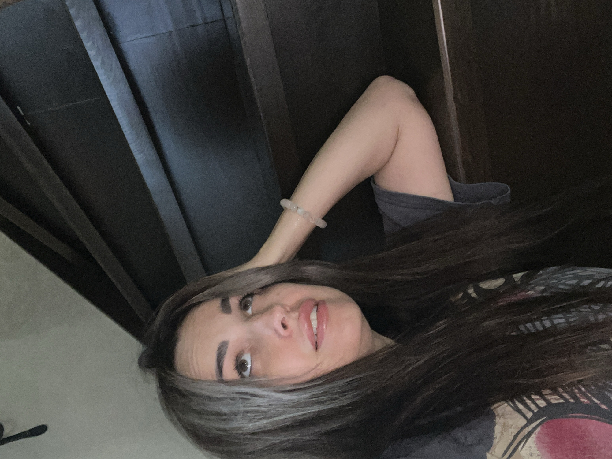 A woman with dark hair and a grey t-shirt is leaning against a dark wooden surface, possibly stairs or shelving. She is looking upwards with her hand on her head and a bracelet on her wrist. The background is dark, with wooden slats visible.