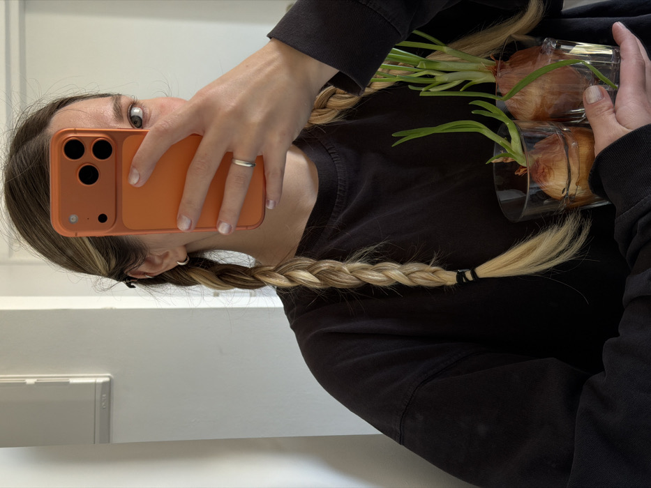 A person is depicted in a self-portrait, taken with an orange-cased smartphone held in their right hand, obscuring most of their face except for the right eye. The individual has long, light-colored hair styled into two braids, secured with black elastic bands, and wears a dark, long-sleeved shirt. A silver hoop earring is visible in the left ear, and a silver ring is on the fourth finger of the right hand. In their left hand, the person holds two clear glass tumblers. Each tumbler contains an onion bulb partially submerged in water, from which several long, vibrant green shoots are actively growing. The background consists of a plain, light-colored interior wall, with a light-colored rectangular fixture visible on the upper left side. The scene is located within the city of Tel-Aviv, Israel.