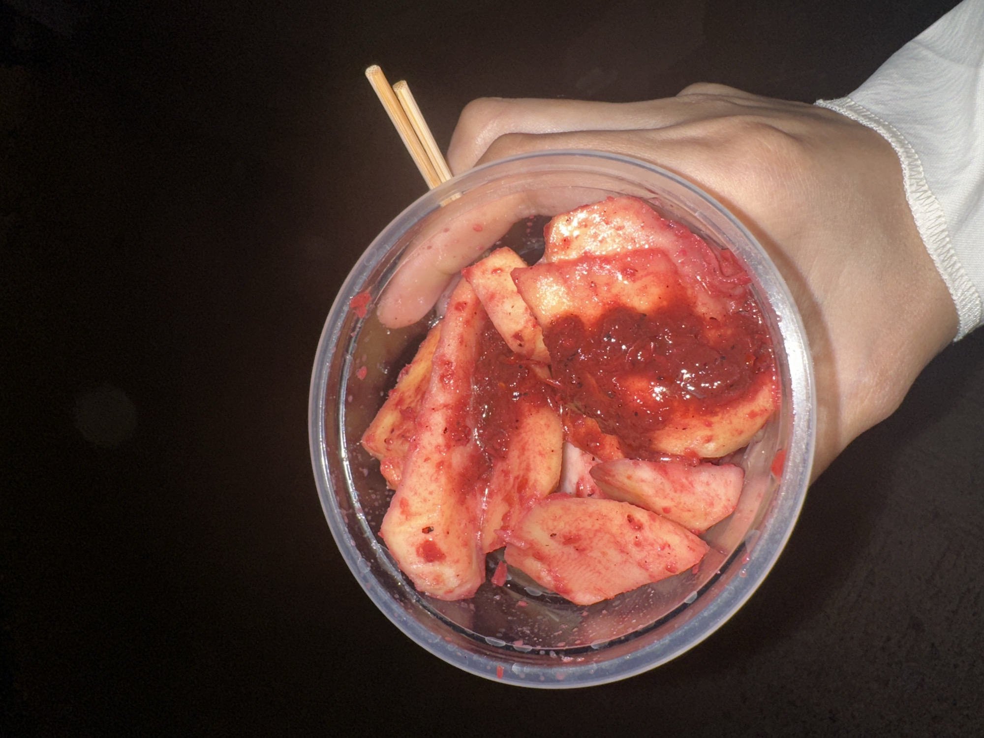 In Davao City, Philippines, a right hand, wearing a white long-sleeved garment with a visible stitched cuff, holds a clear, cylindrical plastic cup. The cup contains multiple irregularly cut pieces of light-colored fruit, likely a variety such as unripe mango or jicama, thoroughly coated in a thick, vibrant red sauce. The sauce appears textured, possibly containing small chili flakes or spices. Two light-colored wooden chopsticks are partially submerged within the fruit and sauce, resting against the inner rim of the cup. The background is uniformly dark and out of focus, indicating low-light conditions, while the subject elements are clearly illuminated.