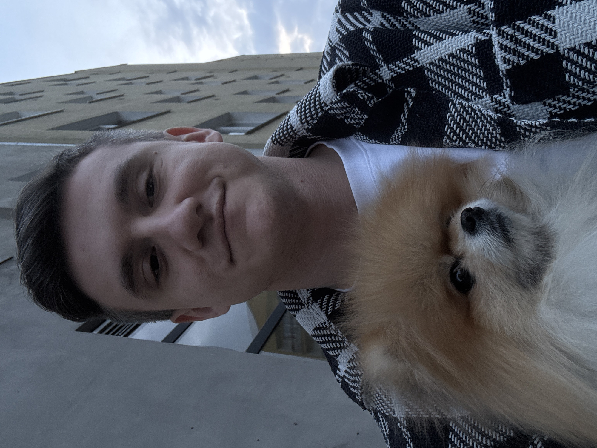 A man is holding a Pomeranian-type dog in front of him, likely taking a selfie. He is wearing a white t-shirt and a black and white checkered jacket or blanket. The man's head and the dog's head are close together. The background includes a tall building with many windows. The lighting suggests it's daytime with some clouds. The location appears to be Warsaw, Poland.