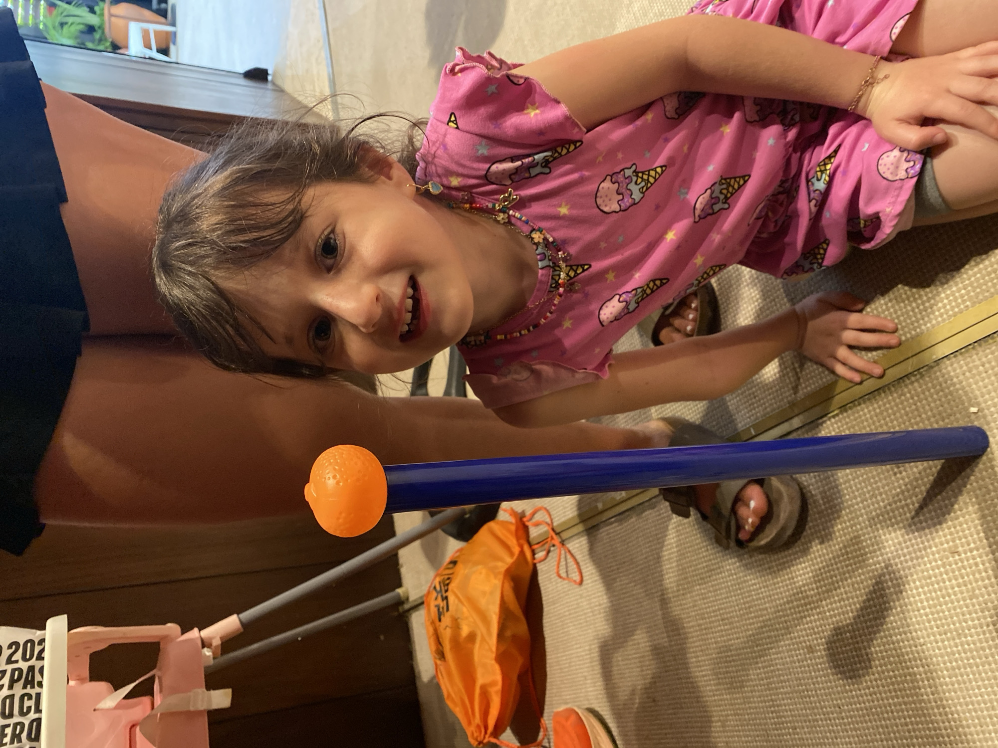 A young girl wearing a pink shirt with ice cream cones and stars is on a beige carpet with a blue tube with an orange ball at the end. There is a pink toy and an orange bag with drawstrings in the image. A woman in a black skirt and brown sandals is partially visible on the left side of the frame. The location is Marina Maroma, Mexico.