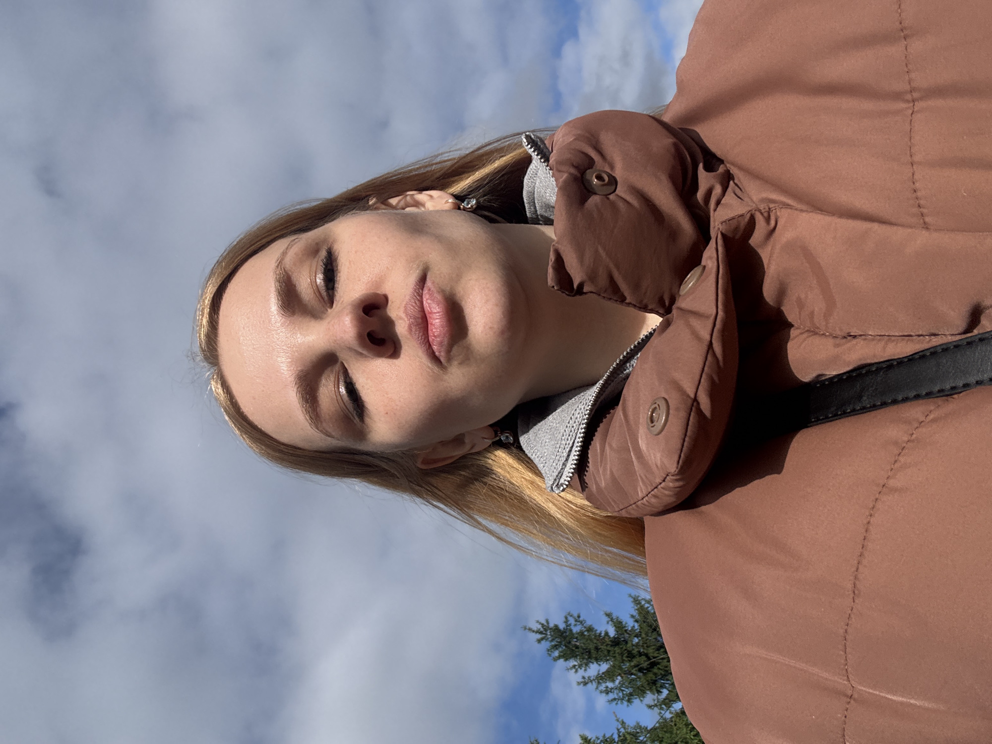A woman with blonde hair is wearing a brown puffer jacket with a gray hood. She is facing the camera at a slight angle, with her eyes partially closed. The background consists of a blue sky with white clouds and the top of a green tree. The location is likely outdoors. The city is not clearly evident in the image.