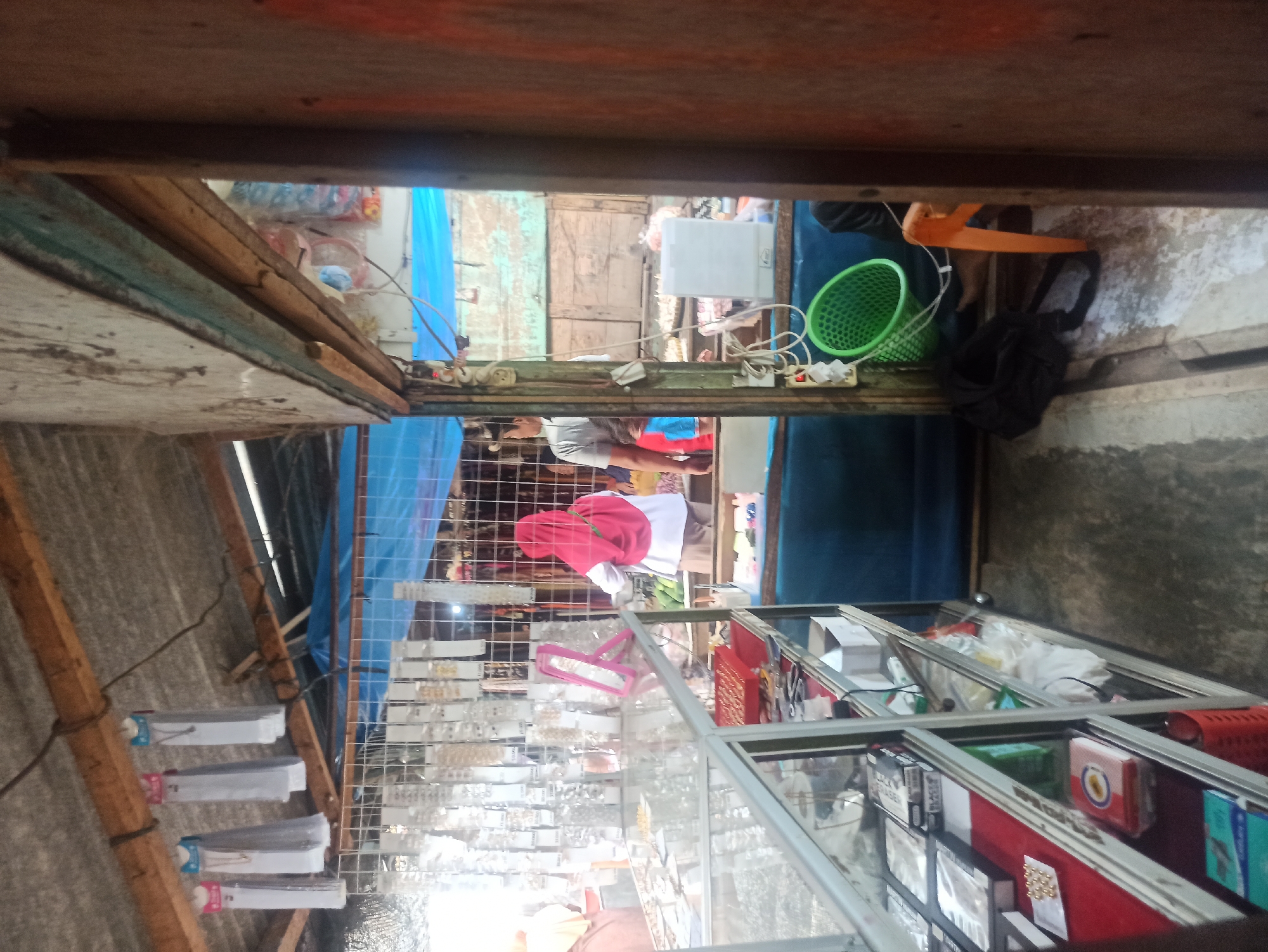 The image depicts a small, open-air market stall in Trisinar, Indonesia. The stall appears to be selling costume jewelry and accessories. A display rack with various items, possibly necklaces and earrings, is visible behind a glass counter. Two individuals are present: one, a person wearing a red hijab and a white top with a pink trim, is interacting with the merchandise. The other individual, partially obscured, is wearing a light-colored shirt and shorts. The stall is constructed with a corrugated metal roof and wooden supports. The environment is somewhat cluttered, with various items, including a green basket and a black bag, scattered around the area. The ground is a rough, possibly concrete, surface.