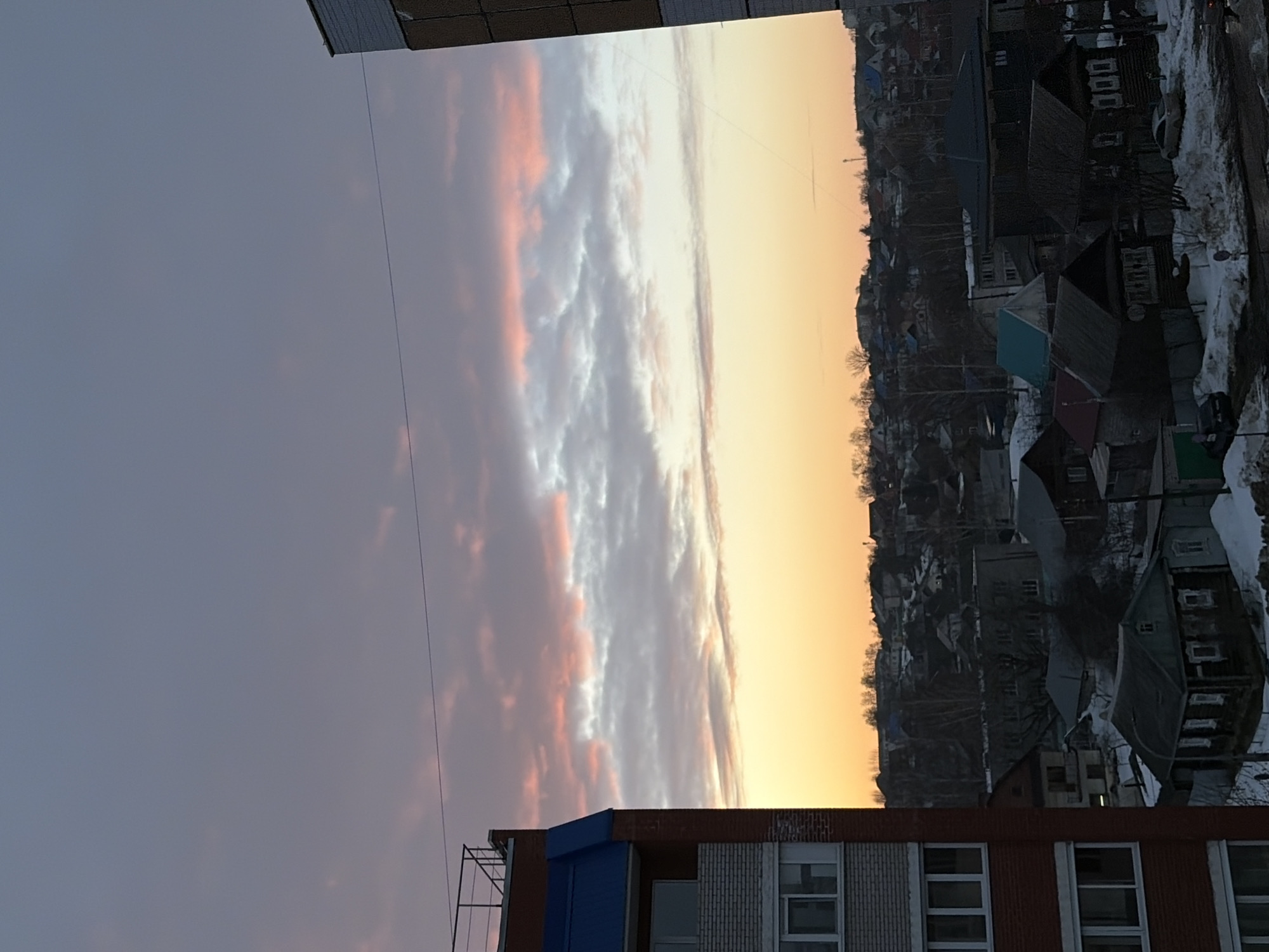 This image displays an urban landscape in Izhevsk, Russia, at twilight. The sky progresses from a deep blue-grey at the top, through strata of pink and grey clouds, to a bright orange and yellow band at the horizon. The left foreground features a partial view of a multi-story building, characterized by brick-red siding, light grey brickwork on lower visible floors, a blue sloped roof section, and a dark metal railing on its rooftop. The right foreground shows a corner of another building with a brown, possibly tiled or paneled, facade. A thin, dark horizontal wire spans the upper middle of the frame, connecting towards the left foreground structure. The midground and background reveal a dense residential area predominantly composed of detached single and two-story houses, many appearing to be older wooden constructions with dark roofs. Patches of dirty snow cover the ground, indicating a cold weather season. Bare deciduous trees are scattered throughout the residential zone. Several parked vehicles are visible on ground-level surfaces in the lower midground. No individuals or specific actions are discernible within the frame.