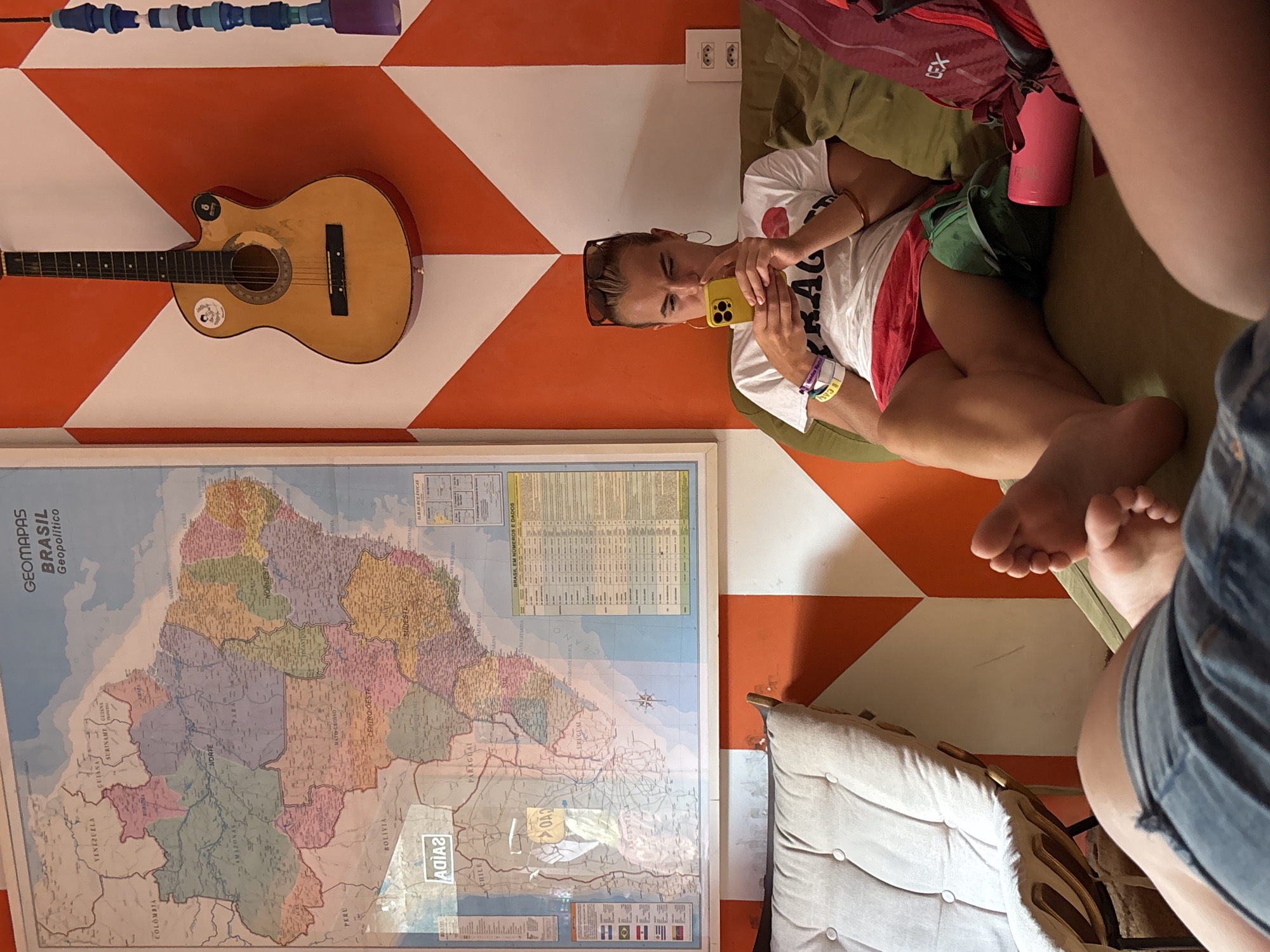 A horizontally-oriented, medium-shot photograph depicts a young woman seated on a plush cushion, intently using a yellow smartphone. She is wearing a white t-shirt with writing on it, red shorts, and has sunglasses on her head. Her feet are visible in the foreground. Next to her is a red backpack and a pink cup. The scene is set against a vibrant orange and white geometric patterned wall.
Acoustic guitar hangs on the wall. Below the guitar is a framed map of Brazil, with a "GEOMAPAS BRASIL Geopolitico" title and labels. Other individuals, only partially visible, flank the right side of the frame.
The location is in São Paulo, Brazil, as evidenced by the map of Brazil displayed on the wall.