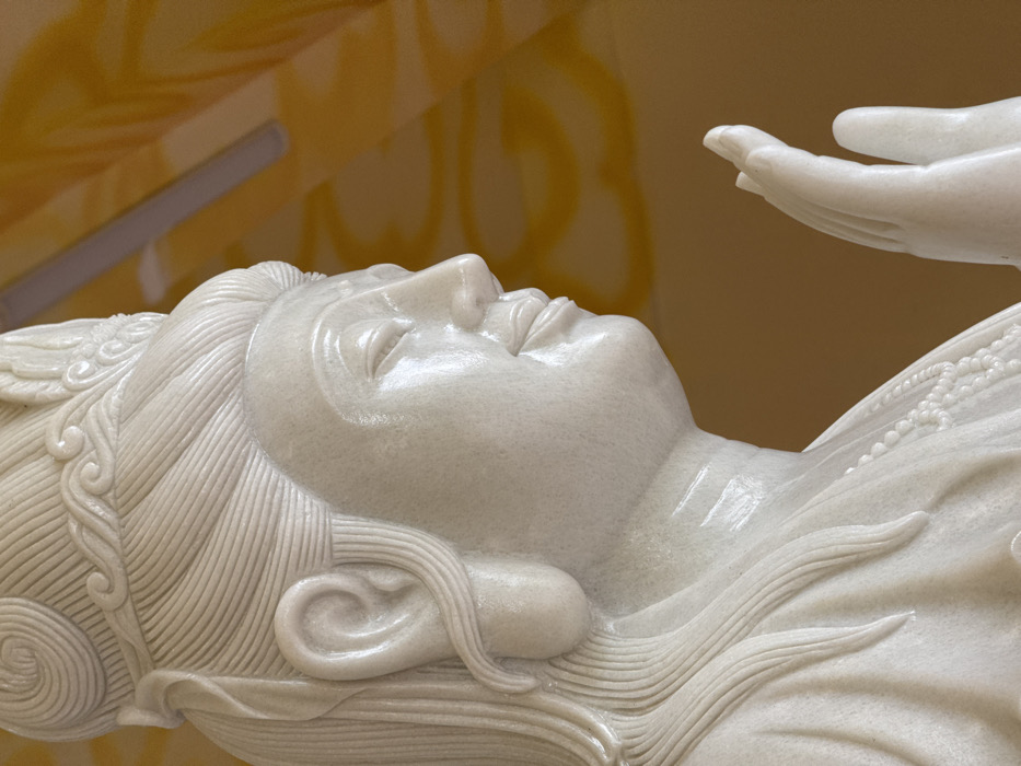 A highly polished white marble statue, depicting a deity or figure, is shown from the upper torso upwards, positioned within an interior setting in the city of Đăk Mar, Vietnam. The statue faces right in profile, featuring meticulously carved hair pulled back and adorned with an ornate crown. Its eyes are depicted as downcast, and a subtle smile is present on its lips. A necklace of sculpted beads rests on the figure's chest. The right hand is raised, palm facing forward with fingers extended and slightly curved. The immediate background consists of a uniformly golden-orange surface, against which a linear fluorescent light fixture is visible in the upper portion of the frame. The statue exhibits a smooth, reflective surface with fine granular textures.