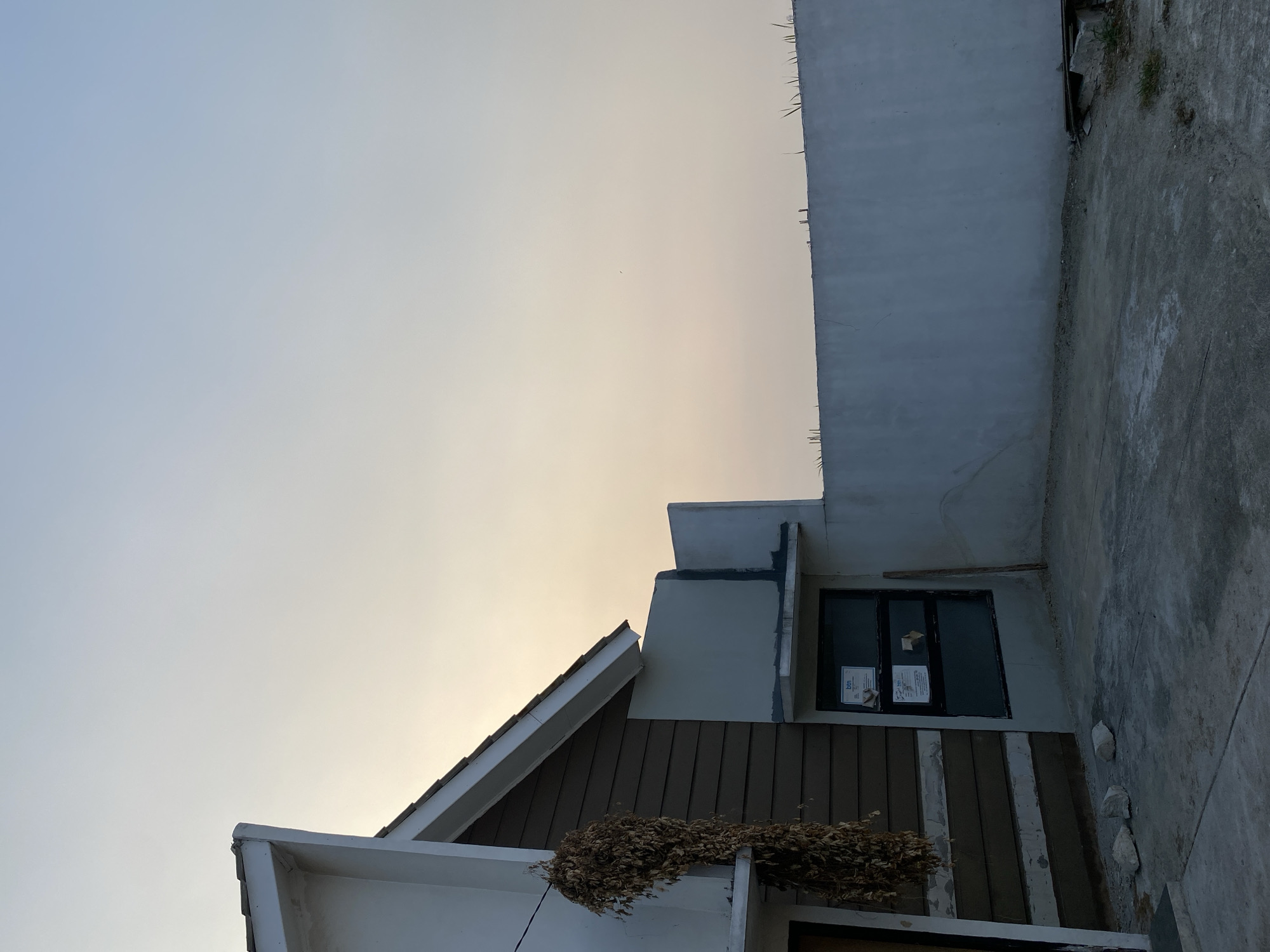 The image depicts the exterior of a modern house under construction, situated in Babelan, Indonesia. The sky is a pale, hazy orange, suggesting either dawn or dusk. On the left, the corner of the house features white siding and brown horizontal panels. A bundle of dried leaves hangs from a wire attached to the house. To the right of this section, a large, dark-framed window is visible, with two white stickers affixed to it. A pale blue concrete wall forms the boundary of the property in the background, with sparse green tufts of grass growing along its top edge. The ground in the foreground is a rough concrete surface, with scattered rocks and construction debris visible. A wooden plank leans against the wall near the window. The overall scene suggests an unfinished or recently completed building in a suburban or semi-rural setting.