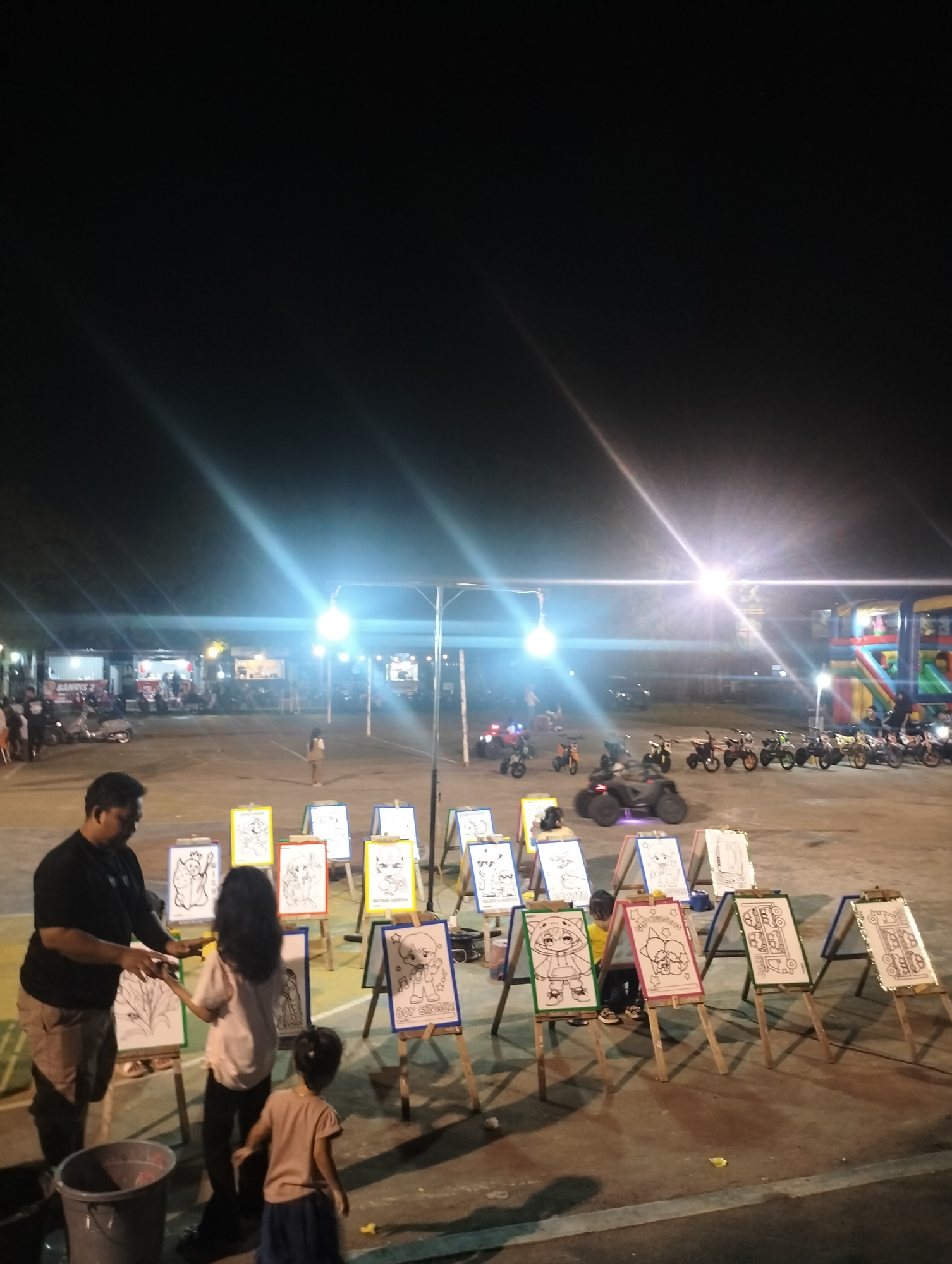 This image captures an outdoor scene at night. Bright overhead lights illuminate the area, casting a glow on the surroundings.
In the foreground, a man is interacting with two children near a series of small easels. Each easel holds a different cartoon drawing. A large bucket sits near the man.
In the background, the scene expands to include a parking lot or open area. Several motorcycles and a four-wheeler are parked in the distance. Behind these vehicles, food stalls are set up. A colorful inflatable structure is located near the edge of the illuminated area.
This image likely depicts a fair or outdoor event taking place in Lhokseumawe, Indonesia. The time of day is night.