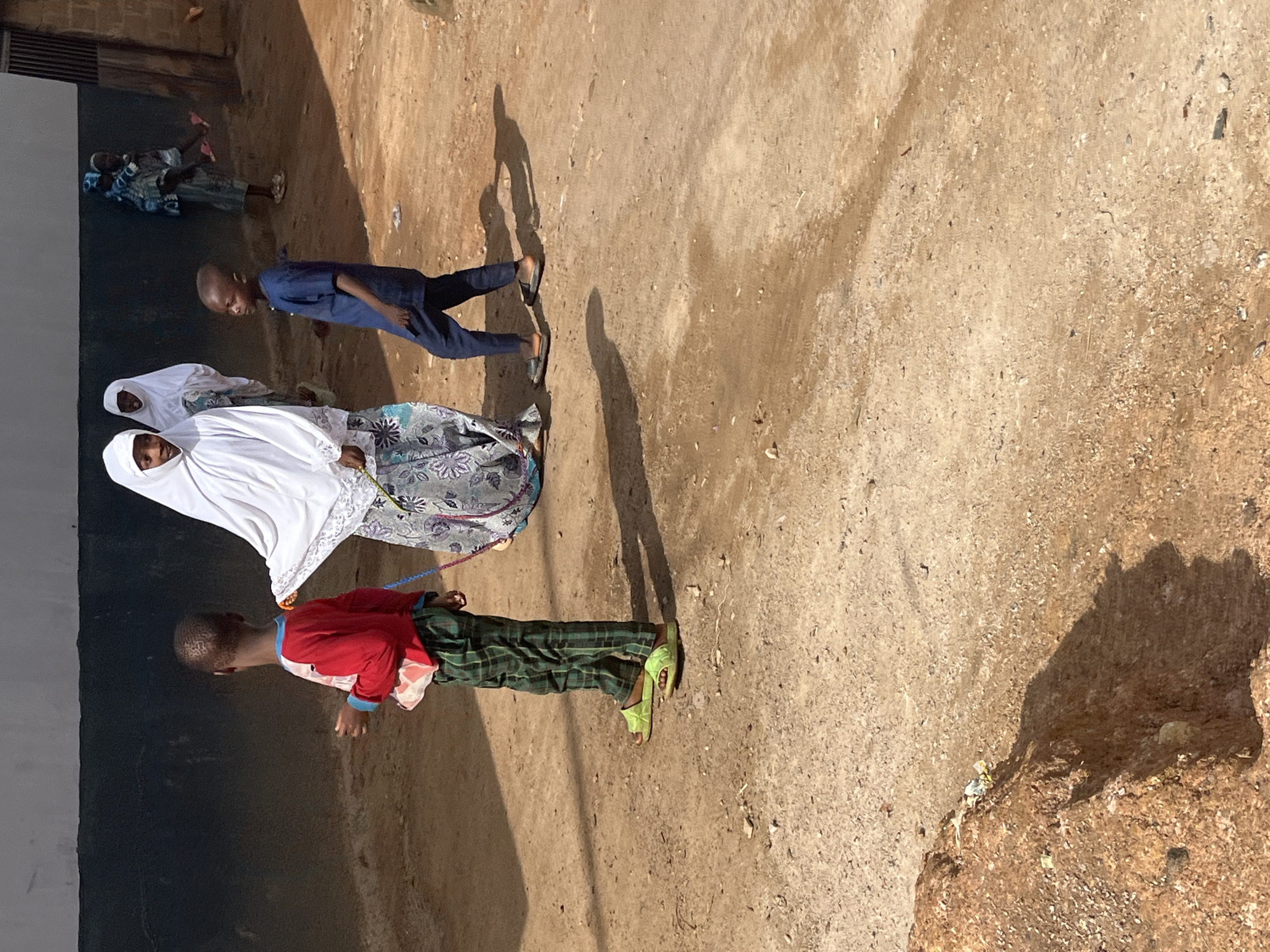 The image captures a group of people standing on a dirt path, likely in Jos, Nigeria. The sun casts strong shadows, suggesting it is mid-day. The ground is a mix of sand and small stones, with some scattered debris. To the left, a dark, rough-textured wall runs along the path, with what appears to be part of a building or structure behind it.
In the foreground, a young boy wearing a red shirt and green plaid pants is visible. He has light skin and short, dark hair. He is looking downwards and appears to be walking or standing. In the middle ground, two women are partially visible. One is wearing a white hijab and a patterned skirt. The other, further back, is also wearing a white hijab and a white garment. Their faces are not clearly discernible. Further back, two more figures, possibly children, are dressed in patterned blue and white clothing.
The overall scene suggests an outdoor gathering or a moment in everyday life in a rural or semi-urban setting. The clothing of the individuals, particularly the women's hijabs and the children's attire, hints at the cultural context of the location. The simplicity of the surroundings, with the dirt path and rough wall, further reinforces this. There is no visible text in the image.