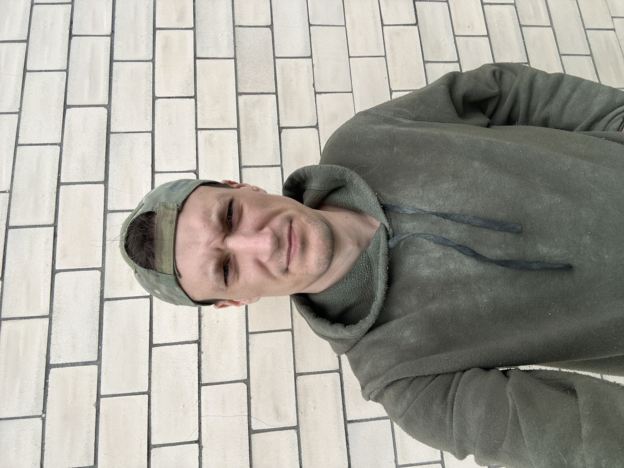 In Makiv, Ukraine, a young man with fair skin, dark hair, and a serious expression wears a dark green hooded sweatshirt and a camouflage baseball cap. He stands in front of a light-colored brick wall, with the man's chest and face filling most of the frame. The man's arms are slightly bent, with his left hand resting on his hip. The brick wall appears to be an exterior wall of a building.