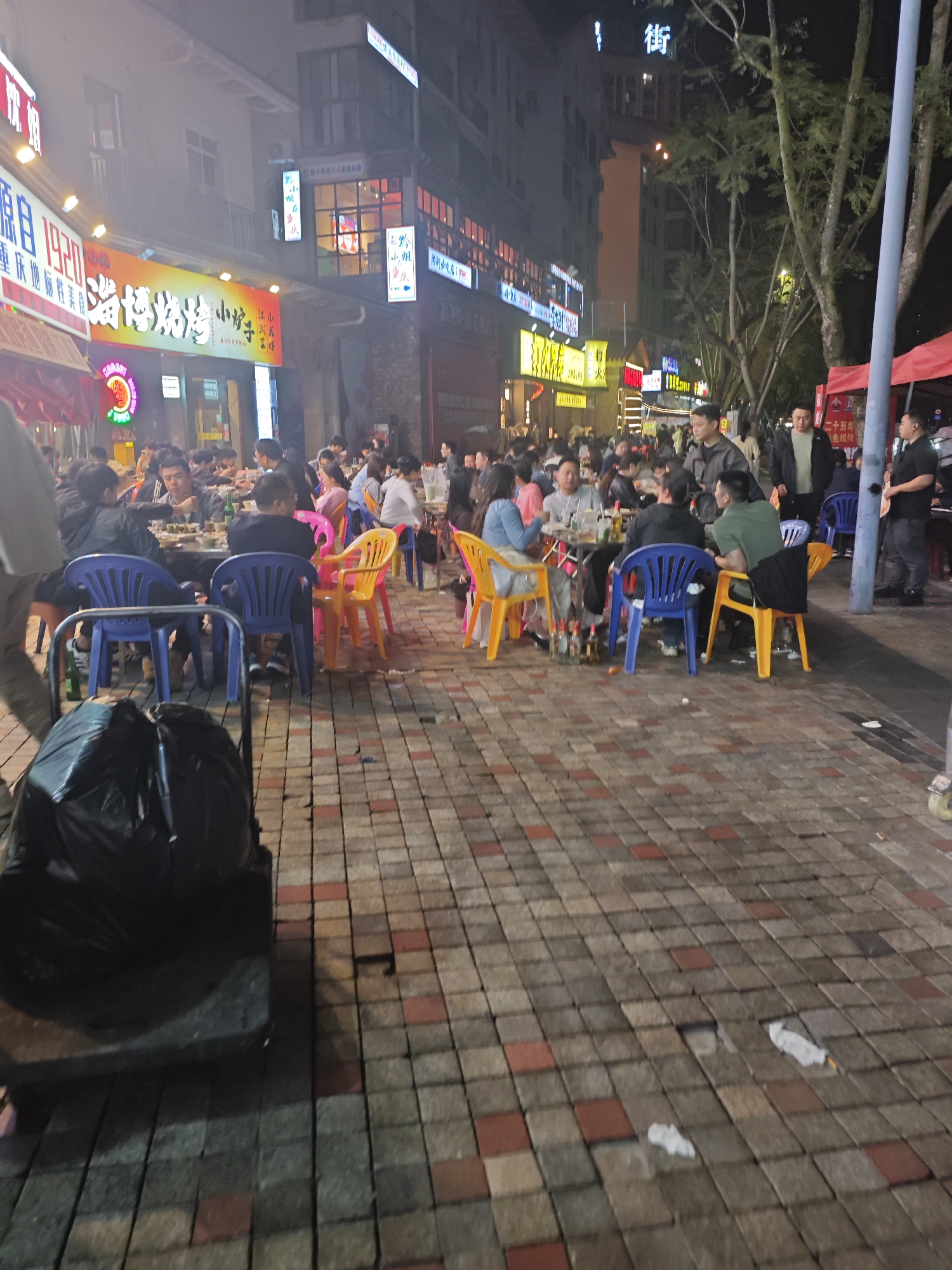 The image shows a nighttime street scene in Chongqing High-tech Zone, China. Numerous people are seated at outdoor tables, eating and socializing. Tables are surrounded by colorful plastic chairs (blue, yellow, pink). Restaurants with bright signage line the street in the background. People are of various ages and are engaged in eating, drinking, and conversation. A trash-filled bag on a wheeled cart is in the foreground. The street is paved with interlocking brick pavers in brown, gray, and red tones. Some litter is scattered on the ground. The light level is relatively high due to the signage and streetlights. Trees are visible in the background.