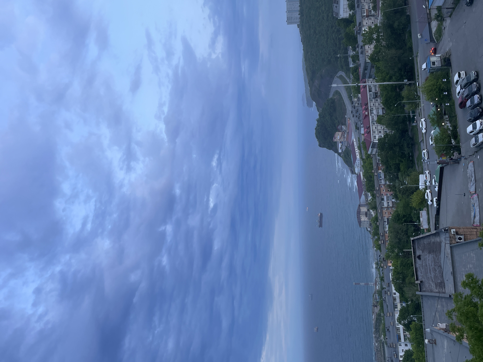 This media file captures a wide panoramic view of a coastal city, likely Vladivostok, Russia, on an overcast day. The image is oriented vertically but depicts a horizontal landscape.
**Subjects:** The upper portion of the image is dominated by a vast, cloudy sky, featuring various shades of blue and gray with textured clouds. Below the sky lies a calm, dark blue sea, stretching towards a hazy horizon. Several large ships, possibly cargo vessels or tankers, are visible on the water in the middle distance.
The coastline is characterized by lush, green, forested hills and mountains that rise directly from the sea. Numerous buildings, predominantly multi-story residential blocks with a mix of red and light-colored roofs, are scattered along the hillsides and closer to the shore. A tall, modern building stands on a hilltop in the distance. Roads and infrastructure wind along the coast and through the urban areas. In the foreground and along the roads, multiple parked cars (mostly white, gray, and black) are visible in a parking lot and along the street. A bright red vehicle is parked near a small building or booth.
**Activity:** The scene is static, capturing the city's environment rather than specific human activity. The ships appear to be anchored or moving slowly in the distance, and the cars are parked.
**Notable Details:** The time of day appears to be daylight, but the heavy cloud cover gives the scene a muted, somewhat dim, and cool-toned appearance, suggesting a cloudy day or possibly early evening. The atmosphere is calm and serene. There are no distinguishable textual elements visible in the image. The combination of hilly terrain, a large body of water, and distinct architectural styles aligns with the specified location context of Vladivostok.