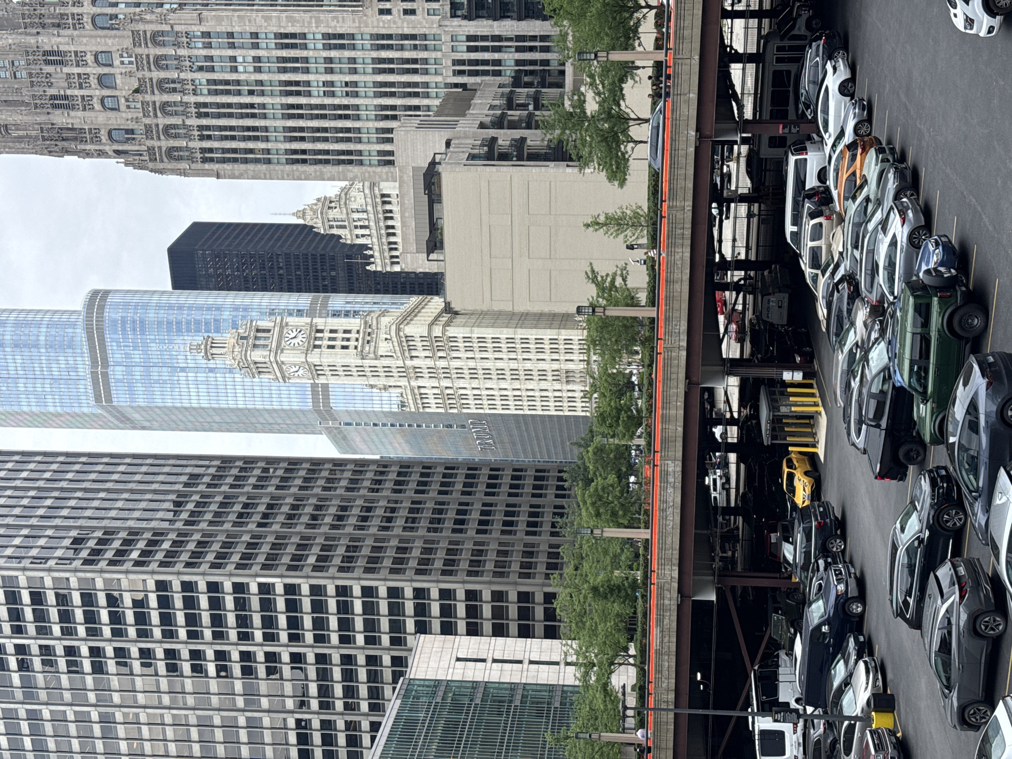 This image captures a view of the Chicago Riverfront with the Trump International Hotel & Tower prominently featured on the right side, identifiable by the "TRUMP" signage. To the left of the Trump Tower are the historic Wrigley Building and the modern skyscrapers of the Chicago skyline. In the foreground, a parking structure is visible with various cars parked, including a yellow sedan and a white van. A pedestrian walkway with trees and streetlights is situated above the parking structure, separating it from the riverfront buildings. Several people are visible walking on the walkway. The sky is overcast, suggesting a cloudy day.