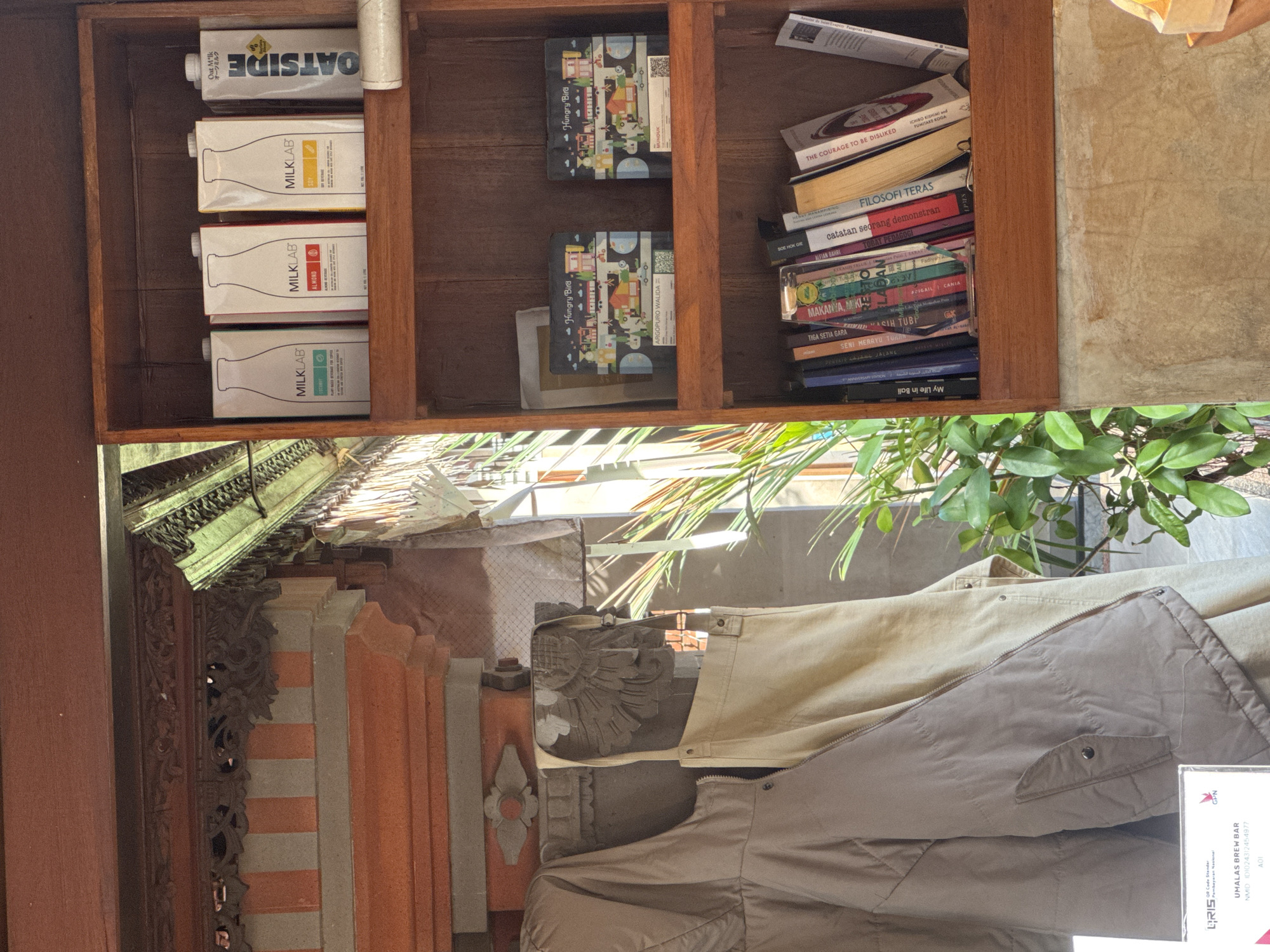 The image depicts an indoor scene with an emphasis on a wooden shelf unit. On the top shelf of the unit, four cartons of milk are visible: MilkLab coconut, MilkLab almond, MilkLab soy, and Oat M'lk Oatside. Below these, on the second shelf, are two bags of coffee, both labeled "Hungry Bird," with one marked "ARGOBURO WALIDA" and the other "MUNDUK." The third shelf contains a collection of books, with visible titles including "My Life in Bali," "Tiga Setia Guna," "MAKANAN AKU," "ILMU TEKNOLOGI," "SEJUTA UMPET," "filosofi teras," "The Courage to be Disliked," and various other books with Indonesian titles. The wooden shelf unit is positioned next to a stone wall with a textured surface. In the foreground, to the left, are hanging garments, possibly jackets or aprons, in neutral tones. A small sign with a QR code and text that includes "UMALAS BREW BAR" and "PEMBAYARAN NASIONAL" is partially visible at the bottom left. Lush green foliage is seen to the right and behind the shelf unit, suggesting an outdoor or semi-outdoor setting. The overall aesthetic suggests a cafe or boutique establishment in a tropical location. Based on the "UMALAS BREW BAR" sign, the location is likely Umalas, a sub-village in Kerobokan, North Kuta, Bali, Indonesia.
