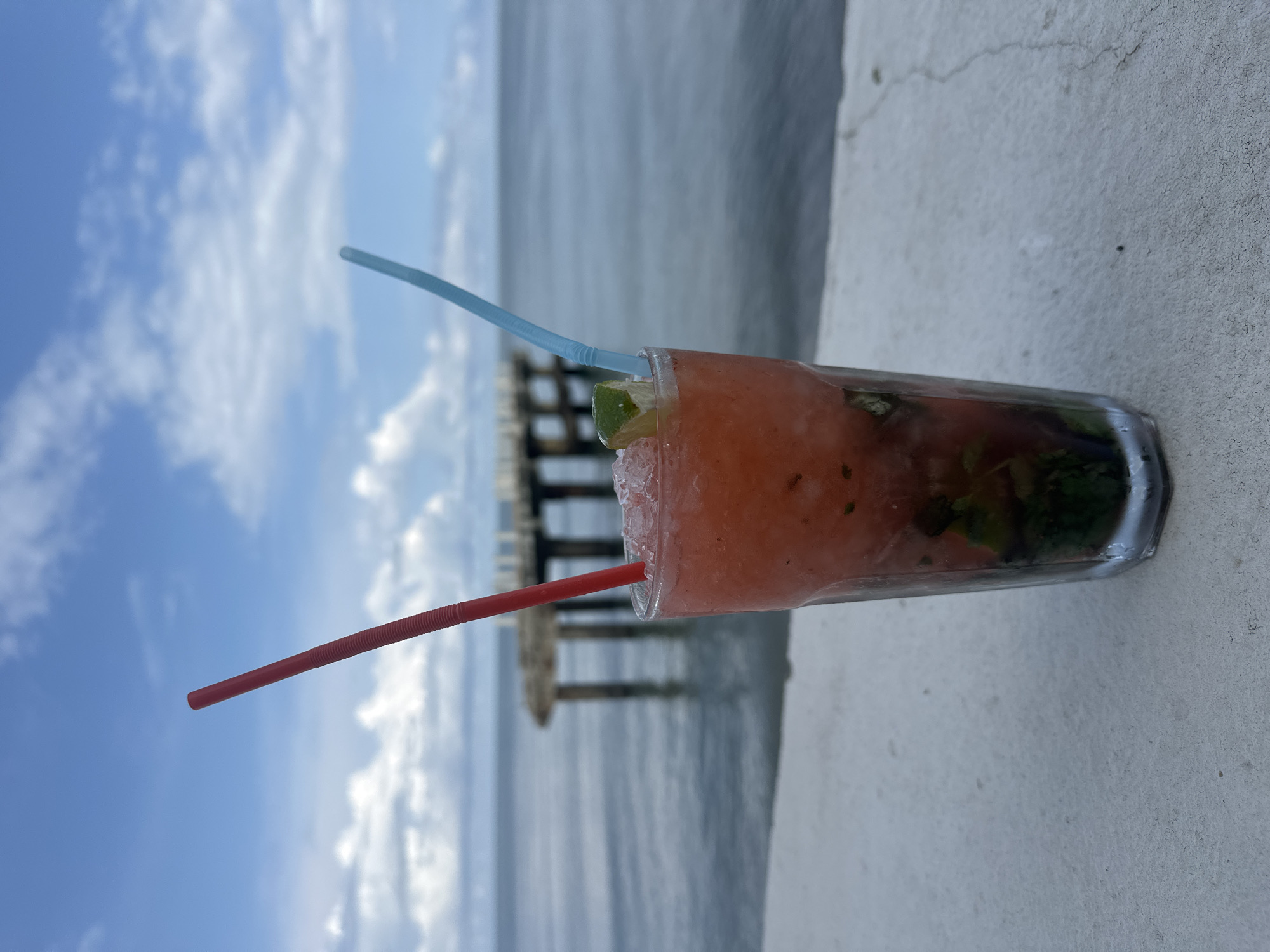 A close-up, eye-level, outdoor shot features a tall glass of a vibrant red, icy beverage garnished with a lime wedge and two straws, one red and one blue, inserted into the drink. The glass is positioned on a light gray, textured surface, possibly a concrete ledge, in the foreground. Beyond the drink, a calm, grayish-blue body of water stretches to the horizon, meeting a sky filled with scattered white clouds against a light blue backdrop. In the middle distance, a weathered pier with several concrete pillars extends into the water. The scene suggests a waterfront location, consistent with the coastal city of Batumi, Georgia, known for its seaside promenade and pier.