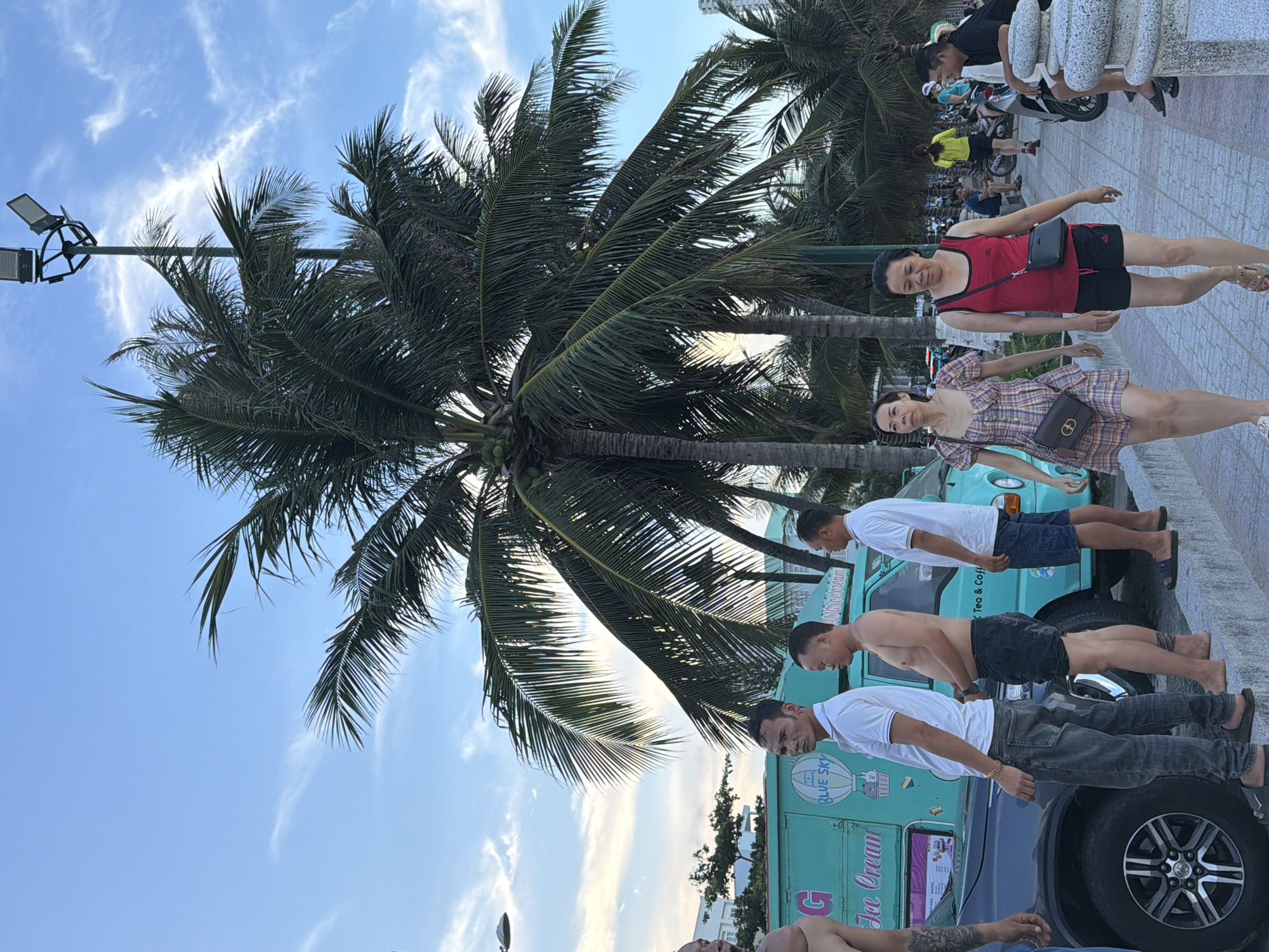 The image shows a street scene in Nha Trang Ward, Vietnam, with a prominent coconut palm tree in the foreground. The sky is blue with some wispy clouds, suggesting it is daytime, likely late afternoon or early evening as the light appears soft.
Several people are visible in the image. In the foreground, on the right, two women are standing. One is wearing a red tank top and black shorts, and the other is in a checkered dress. Behind them, a light blue ice cream truck is parked. The truck has text on its side that reads "BLUE SKY" and "Ice Cream."
Further back, more people are seen, some standing and some on motorbikes. The ground is paved with tiles, and a stone wall is visible. The overall impression is a casual, outdoor gathering on a warm day, possibly near a beach or promenade given the palm tree and the presence of an ice cream truck. The people appear relaxed and are likely enjoying the outdoors. The orientation of the image is landscape, with the top of the image pointing to the right.