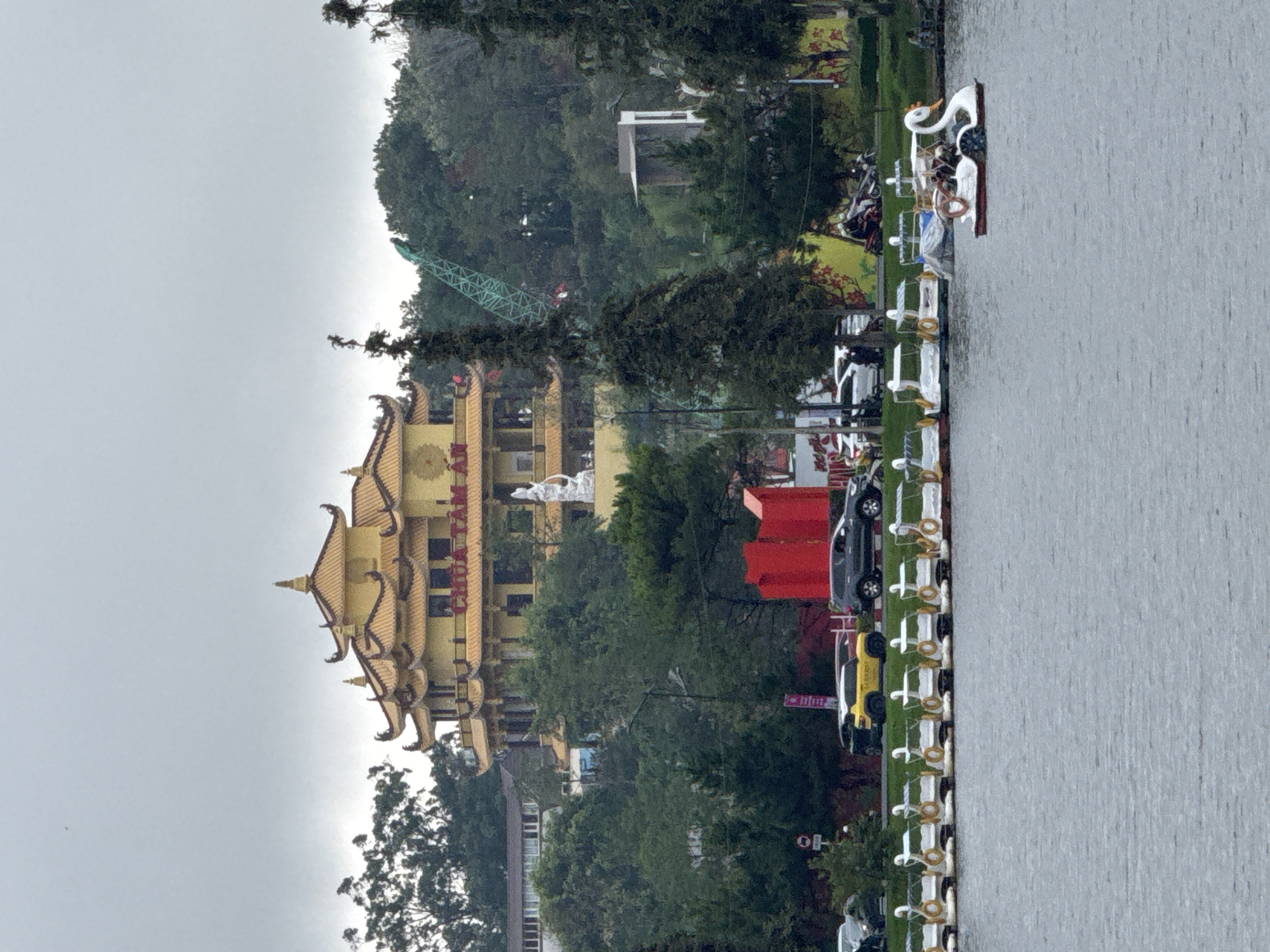 The image captures a scenic lakeside view in Da Lat, Vietnam, under an overcast sky. On the right, a calm body of water, likely a lake, occupies a significant portion of the frame. Along its edge, a white railing with decorative rings or lifebuoys lines a paved promenade. Numerous white swan-shaped paddle boats are neatly docked alongside this railing, with a few other types of boats further down the line. A few individuals are visible near or on these boats, suggesting recreational activity.
On the left side of the image, a lush green hillside rises, densely covered with a variety of trees. A prominent, multi-story yellow building with ornate, curved, tiled roofs stands out against the verdant background. This building, identified by the red text "CHÙA TÂM AN" on its facade, is a pagoda. A white statue, possibly a deity or religious figure, is positioned on a platform directly in front of the pagoda. In the upper-left, partially obscured by trees, a green lattice construction crane is visible on the hillside.
Near the shoreline, behind the white railing, several vehicles including a black and a yellow car are parked. A distinctive red kiosk or structure is also visible in this area. The diffused light indicates a cloudy or overcast day, contributing to the tranquil atmosphere of the scene, which combines natural beauty with cultural architecture.