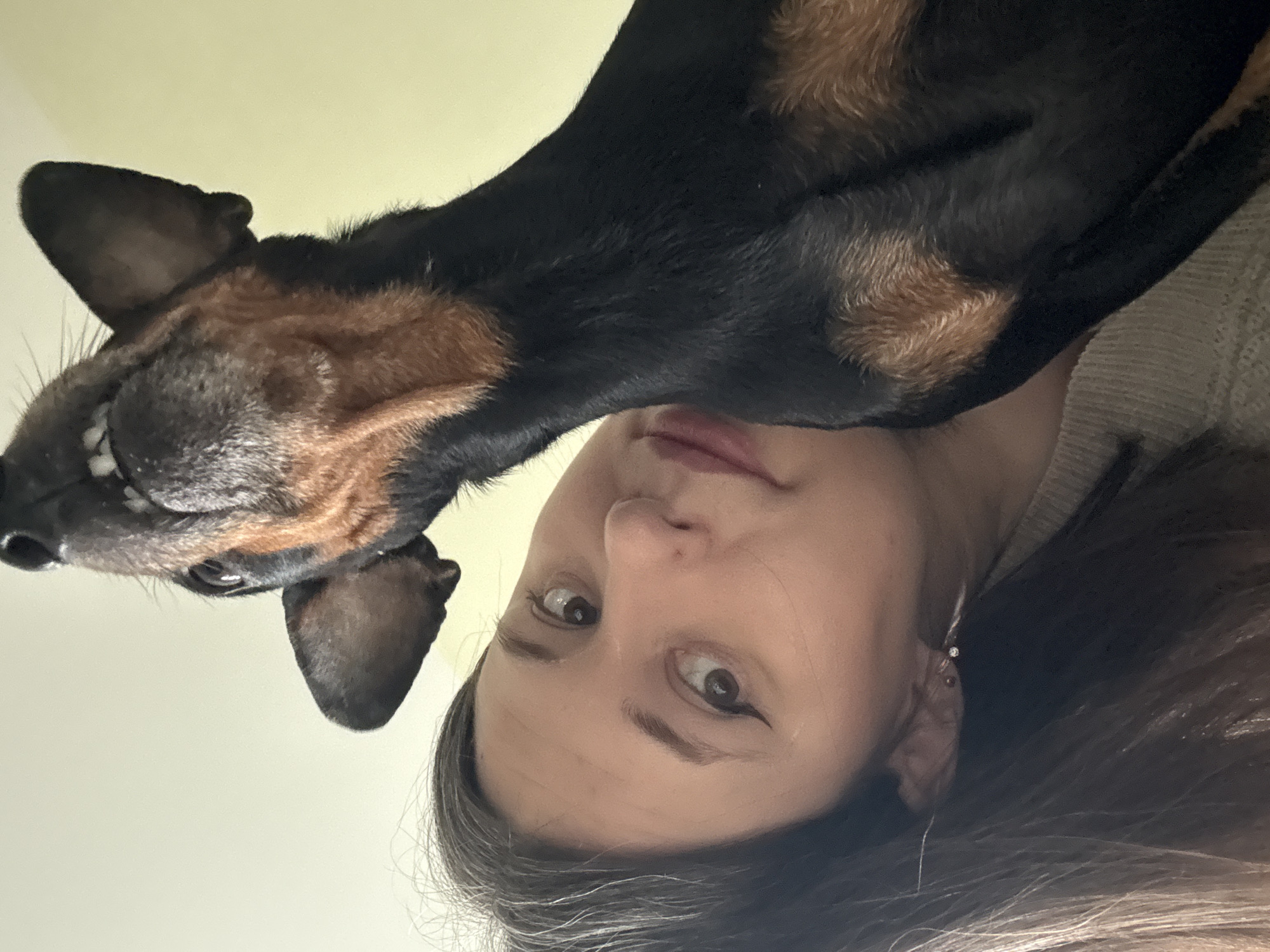 A woman is pictured from a low angle, with a black and brown dog leaning over her face. The woman has brown hair and eyes. The dog appears to be a dachshund mix. The background is a pale yellow color. There is no evidence of the location being in the city of Chaikovsky, Russia.