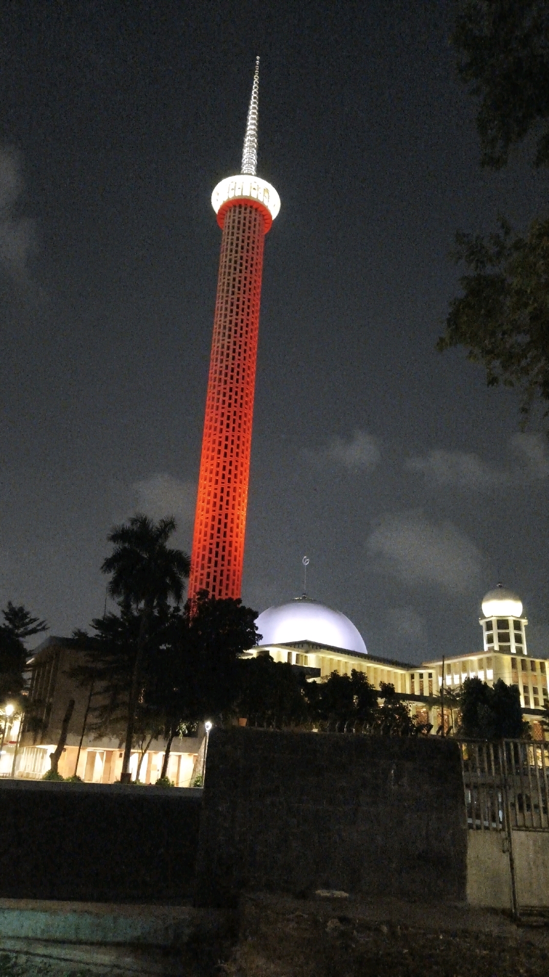 The image depicts the Istiqlal Mosque in Central Jakarta, Indonesia, at night. A tall, slender tower, illuminated with red lights, dominates the center-left of the frame, tapering to a pointed spire at its apex. The tower is surrounded by a white circular band near its top. To the right of the tower, the mosque itself is visible, characterized by a large, white dome and a smaller, square dome atop a building with numerous windows. The mosque and surrounding structures are brightly lit, contrasting with the dark, cloudy night sky. Silhouetted trees, including palm trees, are scattered in the foreground and mid-ground, partially obscuring the lower parts of the buildings. A dark, textured wall and a metal fence are in the immediate foreground, suggesting the viewpoint is from across a street or pathway. The overall scene is one of impressive architectural illumination against a dark night.