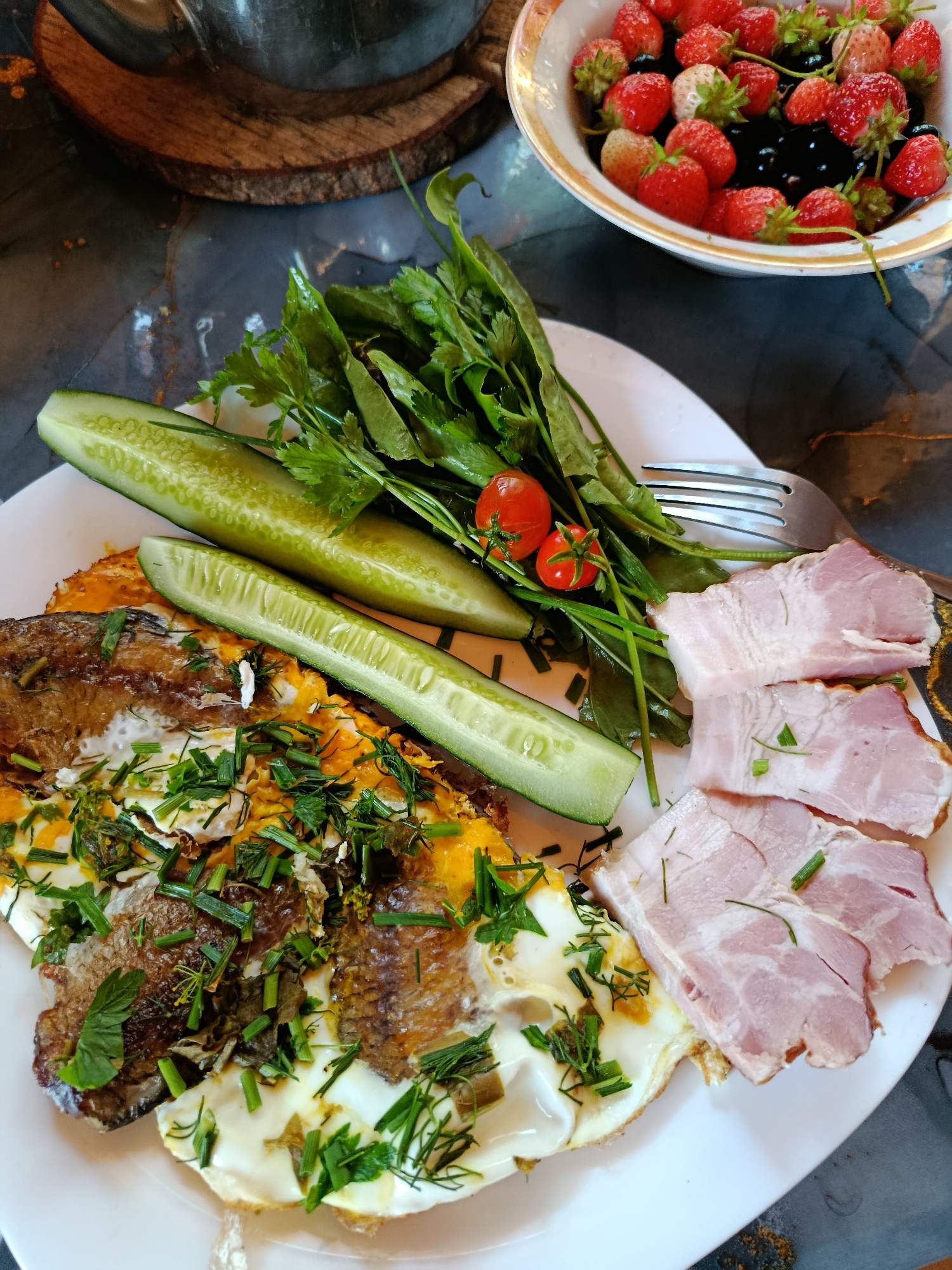 A plate of food is pictured from a top-down perspective. The plate holds fried eggs topped with fish and chopped herbs, two slices of cucumber, a small bunch of green leaves with two cherry tomatoes and a fork, and three slices of ham. In the background, a white bowl is filled with strawberries and dark berries. The food is laid out on a dark, marble-like surface. The scene is well-lit, with the food being the primary focus.