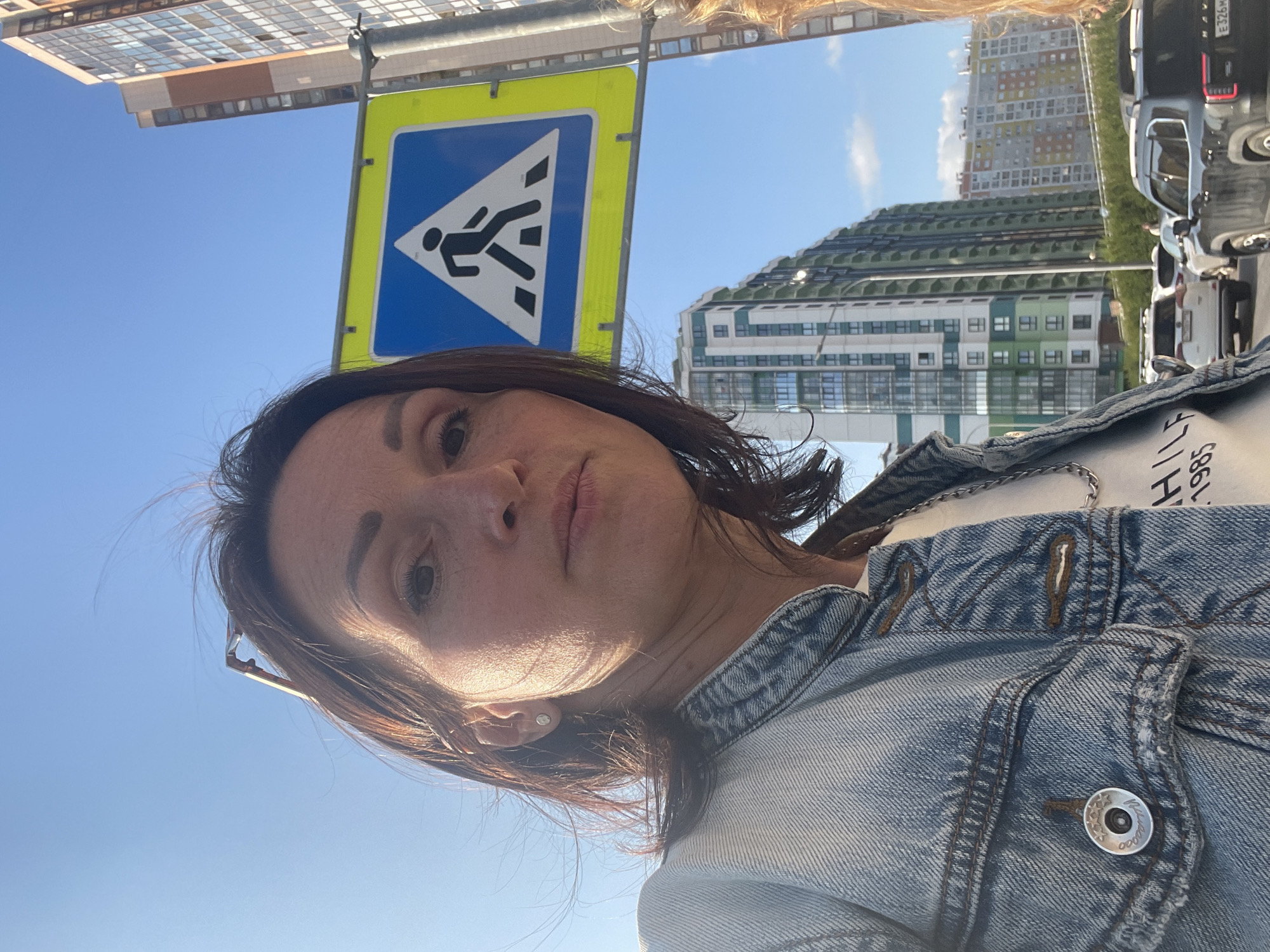 A mid-shot, selfie-style image captures a woman standing on a street in Saint Petersburg, Russia, with a pedestrian crossing sign visible in the background. The woman, appearing to be in her late 30s to early 40s, has brown hair and is wearing a denim jacket over a white t-shirt. She is looking directly at the camera with a neutral expression. Behind her, a bright yellow pedestrian crossing sign is mounted on a pole. In the background, modern apartment buildings with balconies and glass facades rise against a clear blue sky. Several cars are visible on the street, indicating a moderately busy urban environment. The lighting suggests it is daytime, possibly late afternoon, with sunlight casting shadows. The overall scene depicts a casual moment in an urban setting.