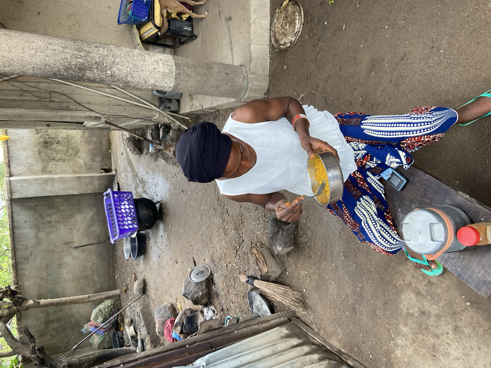A woman wearing a black head covering, white top, and blue patterned pants sits in an outdoor area, presumably in Mararaba, Nigeria. She is holding a metal bowl containing yellow food and using a spoon to eat. She is seated on a wooden bench. To her right is a grey container with a red lid. Around her are various objects including pots, rocks, a broom, and a blue basket. The ground is dirt and concrete. In the background, there is a partially visible dog on a raised platform.