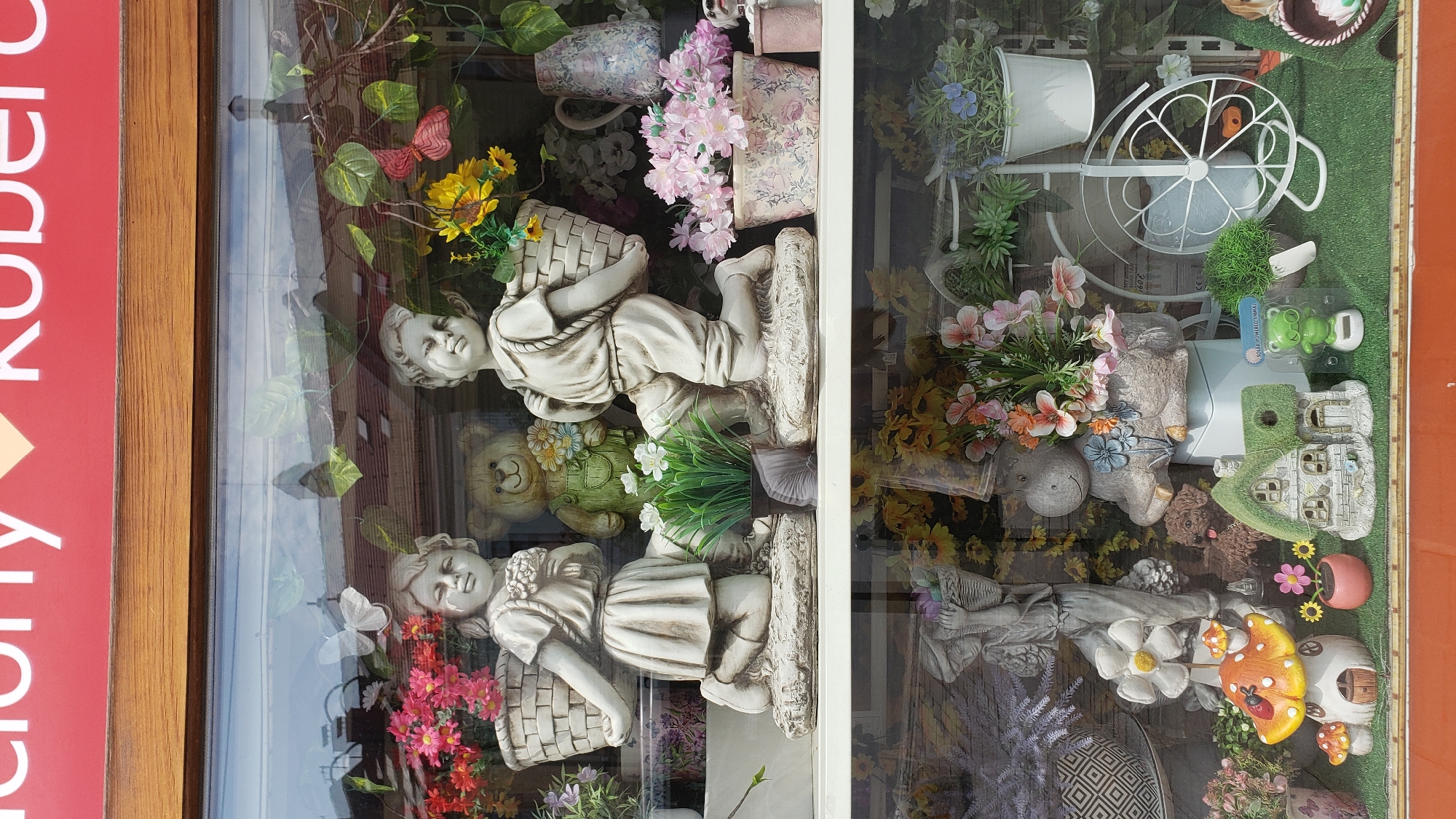 The image depicts a display window of a shop in Prešov, Slovakia. The window showcases various decorative items, primarily garden ornaments and artificial plants. The upper shelf features two stone statues of children carrying baskets, a teddy bear, and floral arrangements. The lower shelf displays a gnome-like mushroom house, a miniature fairy house, a solar-powered frog, and other garden decorations. The overall scene is a well-lit commercial display intended to attract customers.
