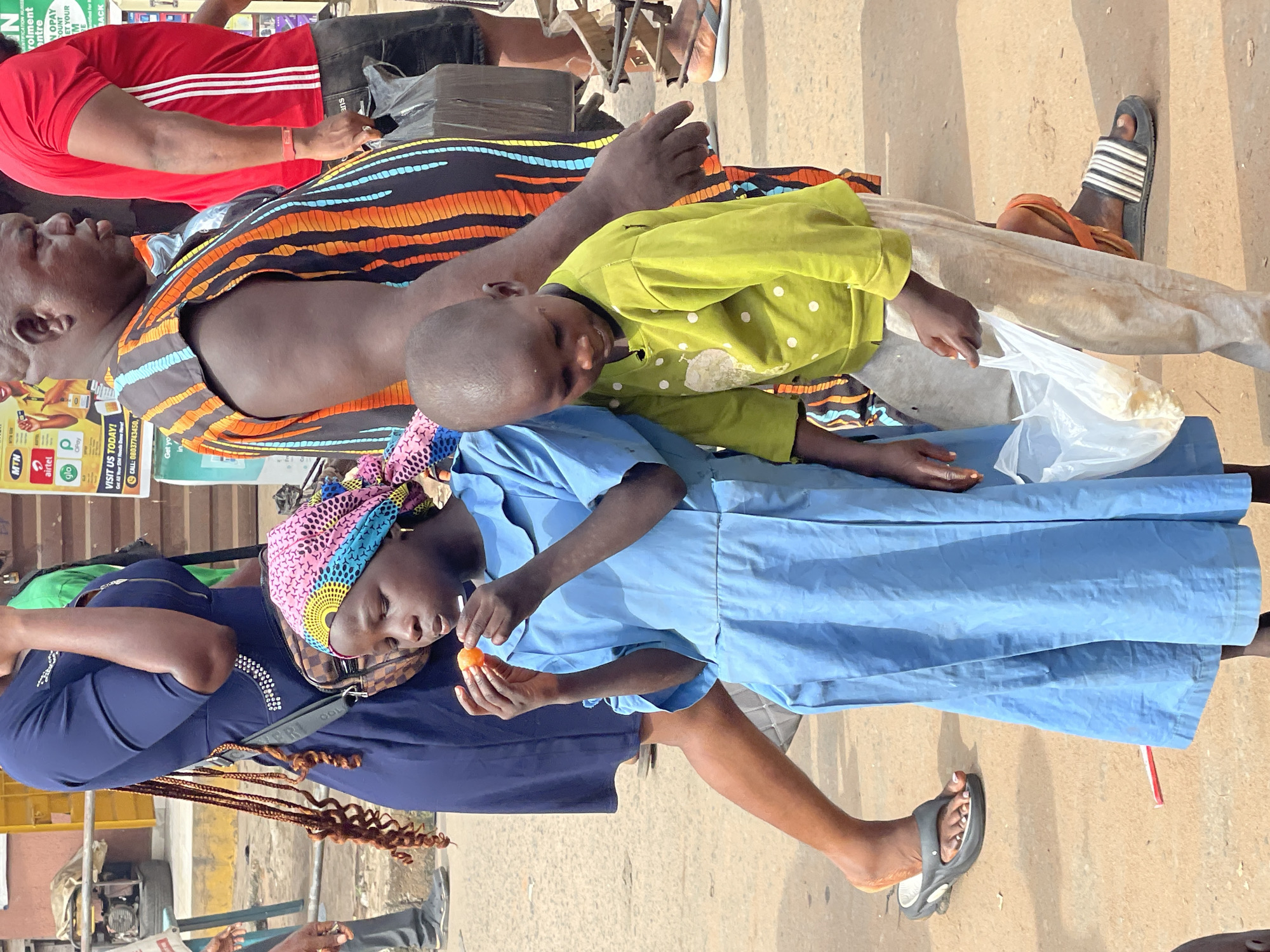 The image depicts a street scene in Nkpor, Nigeria. A young woman in a blue dress and a colorful headscarf is eating an orange fruit. A child in a lime green shirt lies next to her, appearing tired or asleep. Behind them, an adult in an orange and black patterned shirt is visible. A person wearing a red shirt with white stripes is partially visible in the upper left corner of the frame. The ground is concrete, and there are other people and objects blurred in the background, including posters for mobile networks such as MTN, Airtel, and Glo, as well as a poster for OPay. Some sandals are visible on the people's feet. There appears to be a vendor selling products.