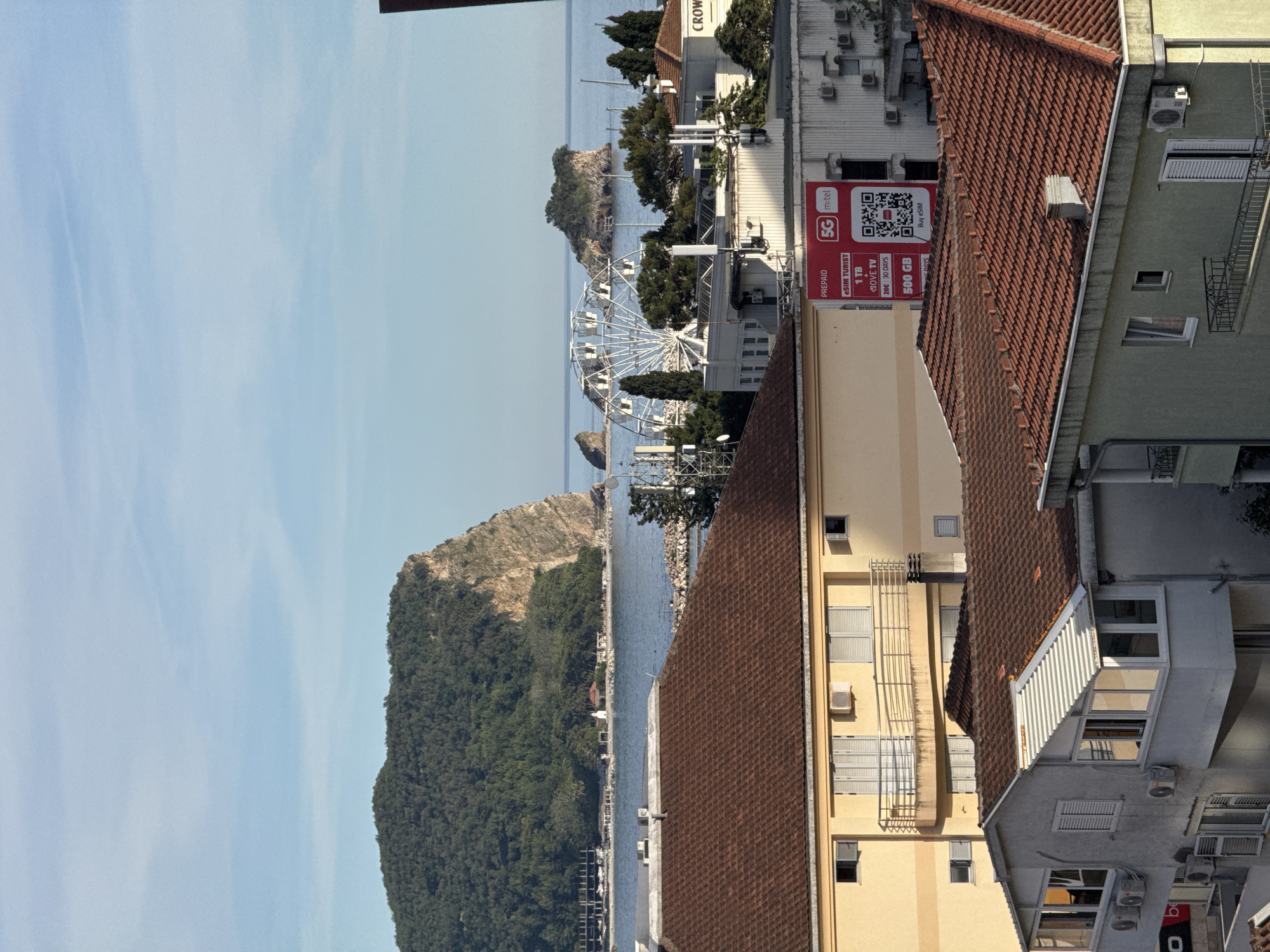 The image shows a view of the city of Budva, Montenegro, from a high vantage point. In the foreground are the tiled roofs of several buildings, with a yellow building on the left and a grey building on the right. The yellow building has several windows and balconies, and appears to be a residential or commercial property. The grey building has a sunroom with large windows. In the middle ground, there are more buildings, including one with a large red billboard advertising "5G m:tel" data plans. Behind these buildings, the Adriatic Sea is visible, with a Ferris wheel and several small islands. In the background, a large, green, tree-covered mountain juts out into the sea. The sky is a clear blue with some wispy clouds. The overall impression is a scenic coastal city with a mix of modern and older architecture.