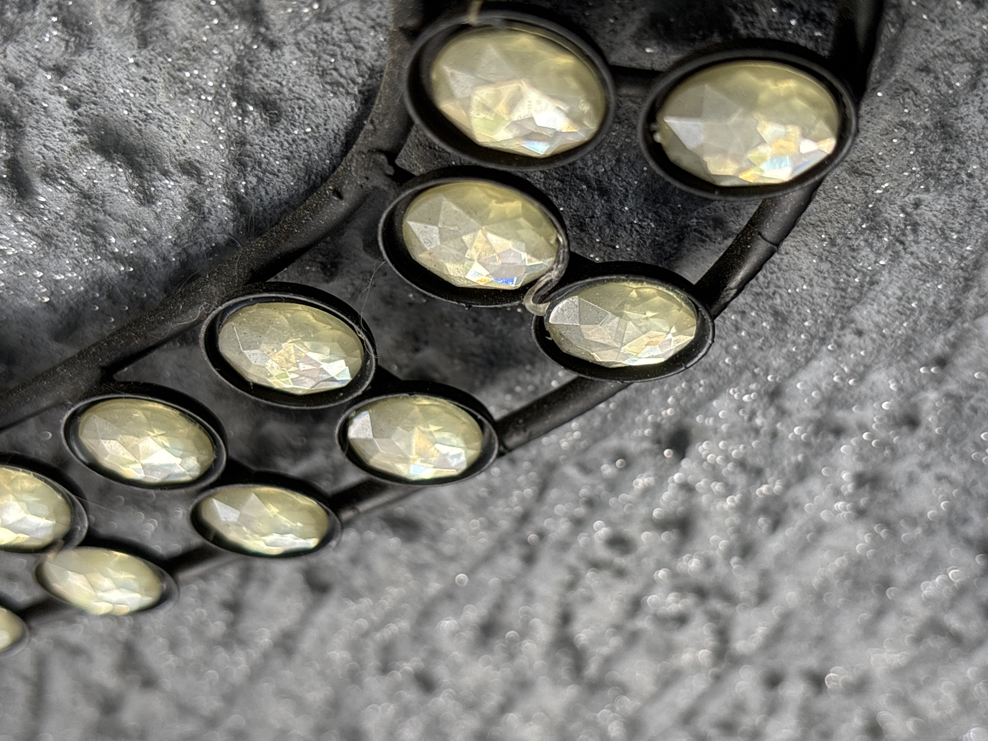 A close-up photograph displays a segment of a decorative object, featuring a series of eleven visible oval-shaped, faceted gemstones or crystals. These gemstones, primarily yellowish-white with iridescent reflections, are individually set within dark, possibly metallic, bezels. The bezels are attached to a curved, dark, thin band or wire, which traverses the frame from the upper left to the lower right. This structure is positioned against a textured, dark gray surface that is uneven and liberally coated with fine, reflective, silvery glitter. The background on the left side of the image exhibits a shallower depth of field, causing the glitter to appear as diffused bokeh. The location is Unknown, Unknown.