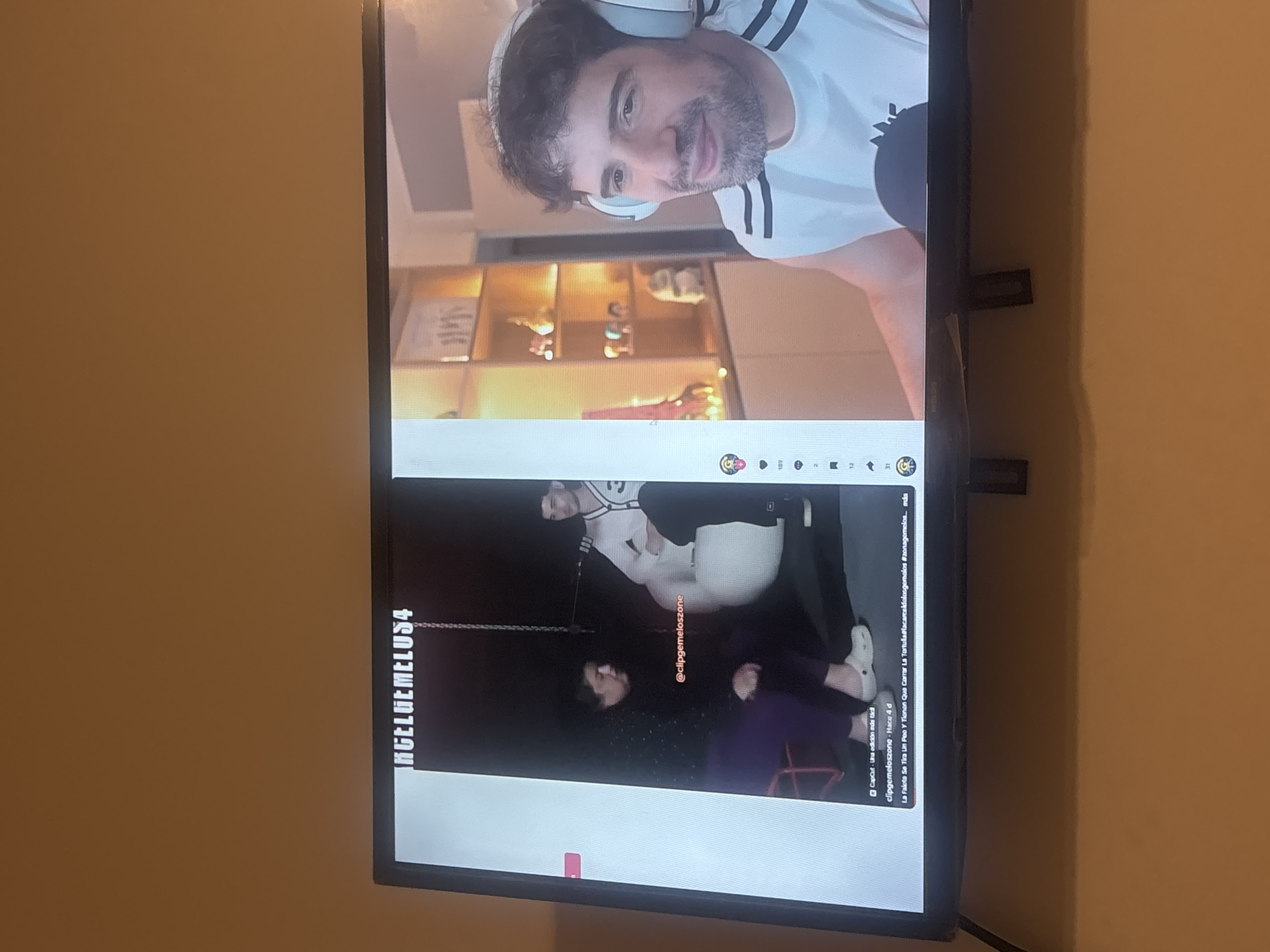 A television displays a split-screen video. The top portion shows a man with a beard and headphones, wearing a white shirt with black stripes. The lower portion depicts a dark scene with two individuals, presumably involved in a TikTok-style skit. Text overlays and usernames are visible on the screen, with a chain hanging in the dark scene. The TV is mounted on a wall.