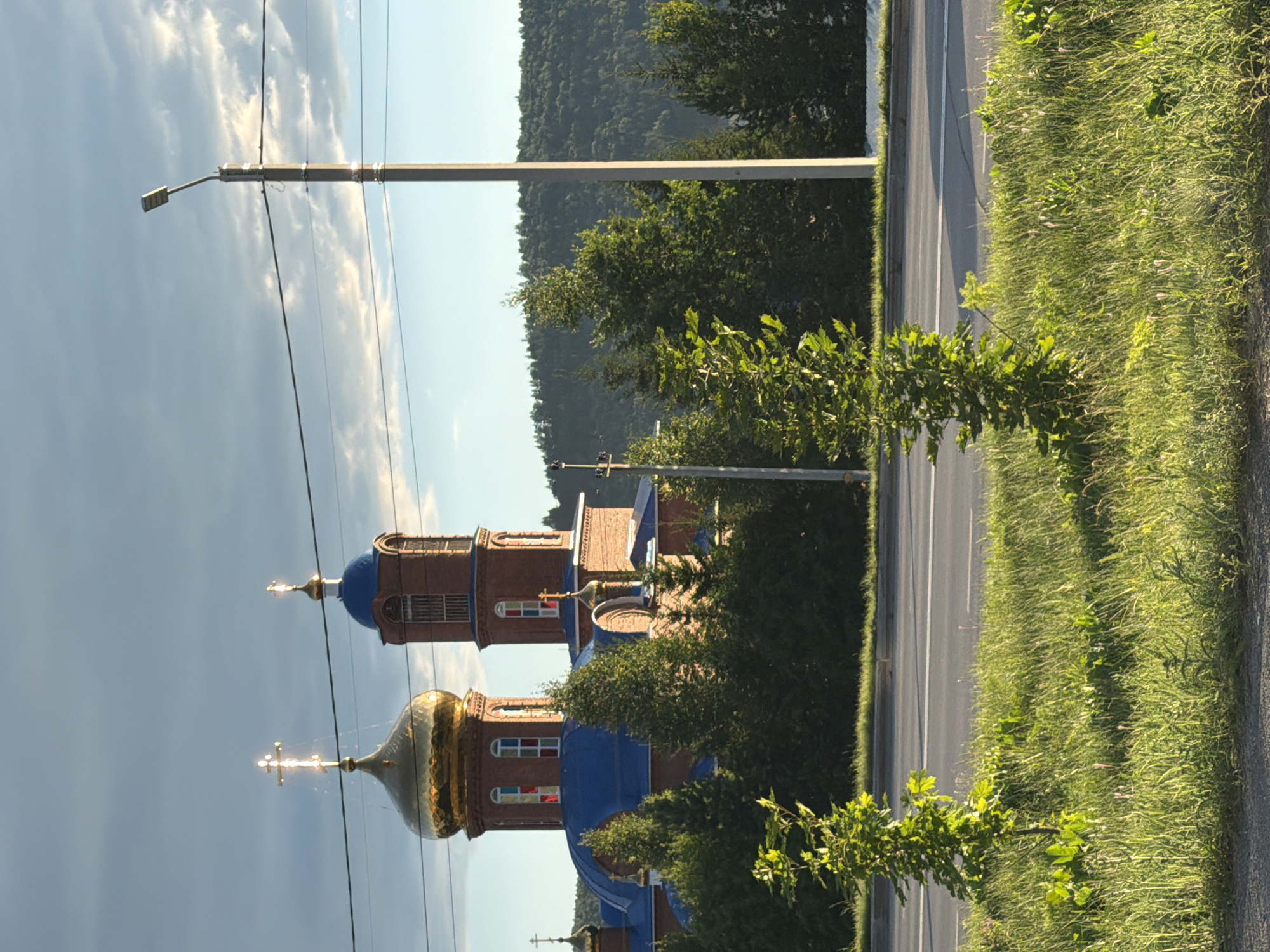 An exterior, eye-level, vertical, wide shot captures a brick Orthodox church with a golden dome and a blue roof, set against a backdrop of a tree-covered hill and a cloudy sky. The church's golden dome is illuminated by the sun, reflecting light. In the foreground, a paved road runs horizontally, bordered by a grassy verge with several young trees. A tall light post stands to the right of the church. The scene appears to be in a suburban or rural setting, with no people or vehicles visible. The time of day seems to be late afternoon or early evening due to the quality of light.