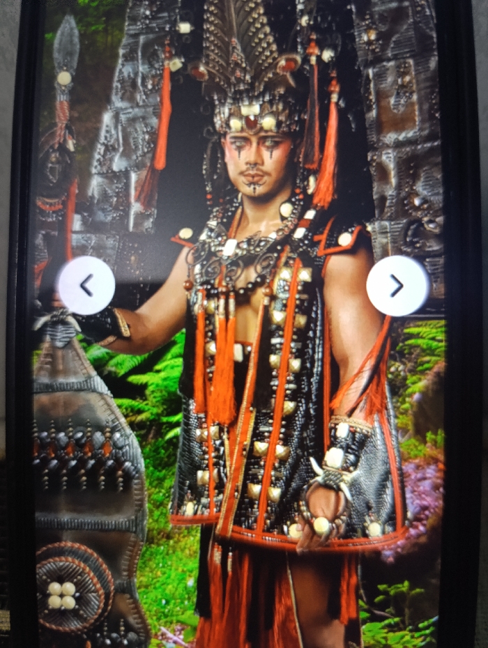 The image shows a man in a traditional costume with a large headdress. The man has dark hair and a beard, he is wearing a long, black and red robe with a dark-colored belt. He is also wearing a beaded necklace and earrings. The man is standing in a green, jungle-like environment. The background features a wall of grey, textured stone with a large, decorative brown shield to the man's left side. The man has a small, white object in his hand. The man is looking at the camera and appears to be in a theatrical performance. The background shows green foliage. The man has a serious facial expression with a dark, decorative mark across his eyes. The image is taken in the Kecamatan Arjasa region of Indonesia. The coordinates for the image are -7.779 latitude, 114.135 longitude. The man is wearing a headdress, the top of which is adorned with many, small, black feathers. The headdress has a red string extending from the top, which is tied behind his head. There is a white circular object in the headdress, positioned above his forehead. There are white, yellow, and red tassels on the headdress, which extend behind the man's back. The man is wearing a black and red vest with various beads and small decorative objects sewn into the vest. The vest has a gold, decorative border. The man is also wearing a dark, beaded bracelet on his left wrist, and he is holding a white object in his right hand. The man's attire and facial markings suggest he may be a member of a tribal group. The image appears to be a photograph or screenshot from a theatrical performance.