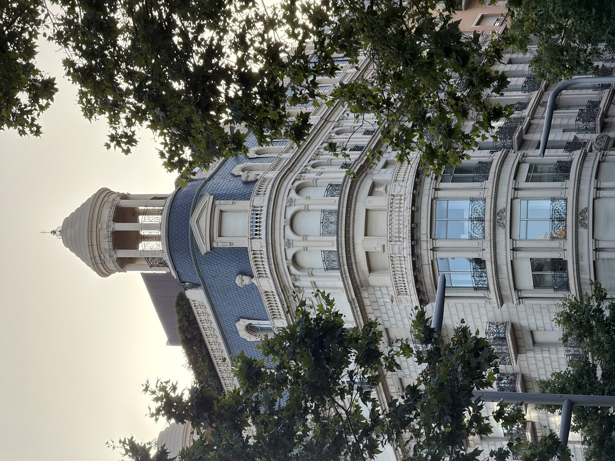 This is a ground-level, exterior, eye-level shot of a building with a rounded corner, possibly in Barcelona, Spain. The building is made of stone and features a dark blue tiled roof with a small turret on top. The building has multiple windows with decorative iron balconies. Trees with green leaves partially obscure the view of the building. The sky is visible in the background, and it appears to be either sunrise or sunset.