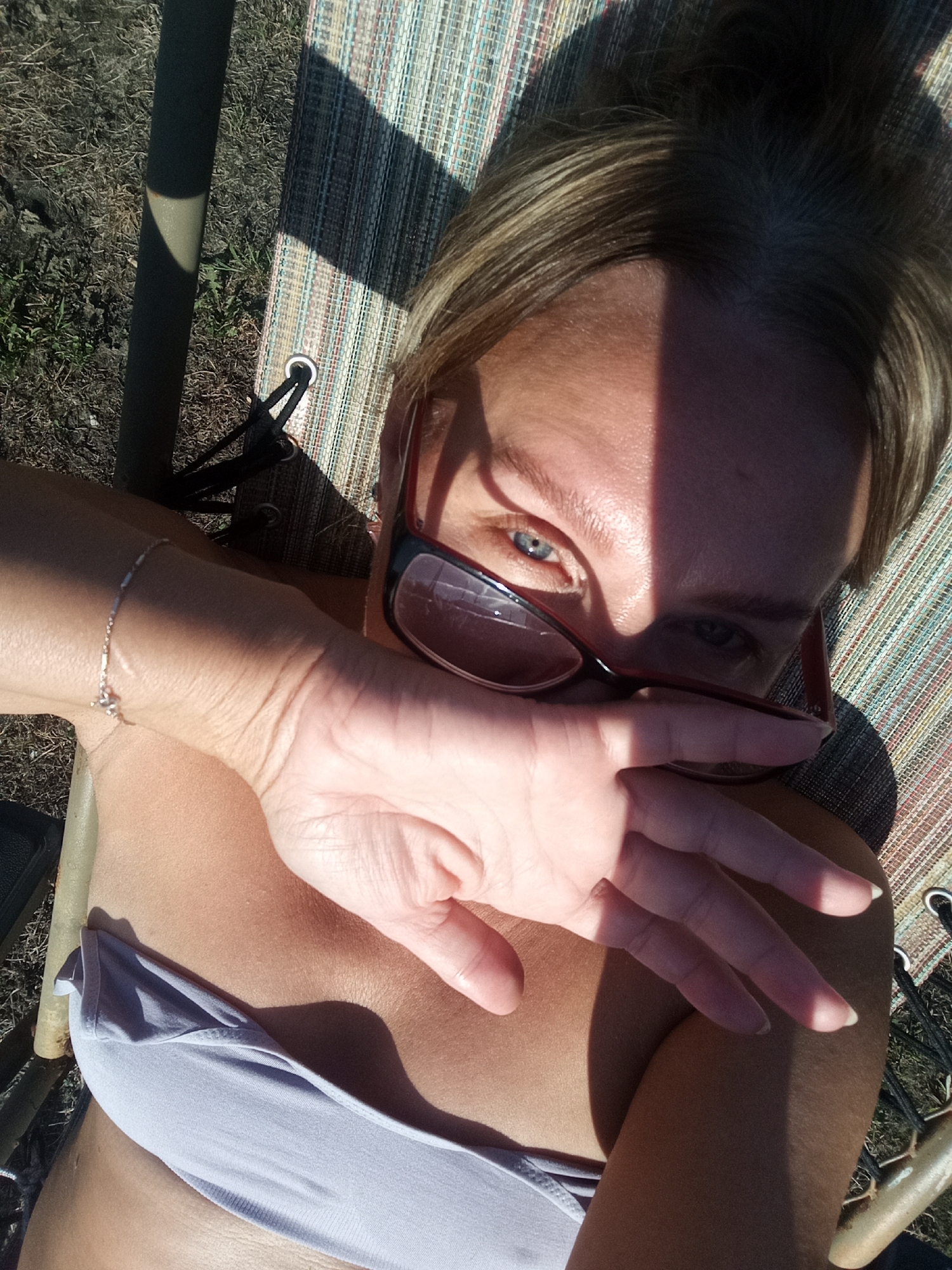 The image depicts a person lying on a lounge chair outdoors, with the camera positioned above and looking down. The subject is partially obscured by their own hand, which is covering their mouth and lower face. They are wearing a light-colored bikini top. Their right arm, adorned with a delicate bracelet, is extended. Sunglasses with red frames are resting on their nose. The background shows dry grass and the textured fabric of the lounge chair, with sunlight casting strong shadows. The image appears to have been taken in a rural or outdoor setting, as indicated by the natural surroundings. There is no identifiable information within the image to suggest the specific location in Novopokrovka, Russia.