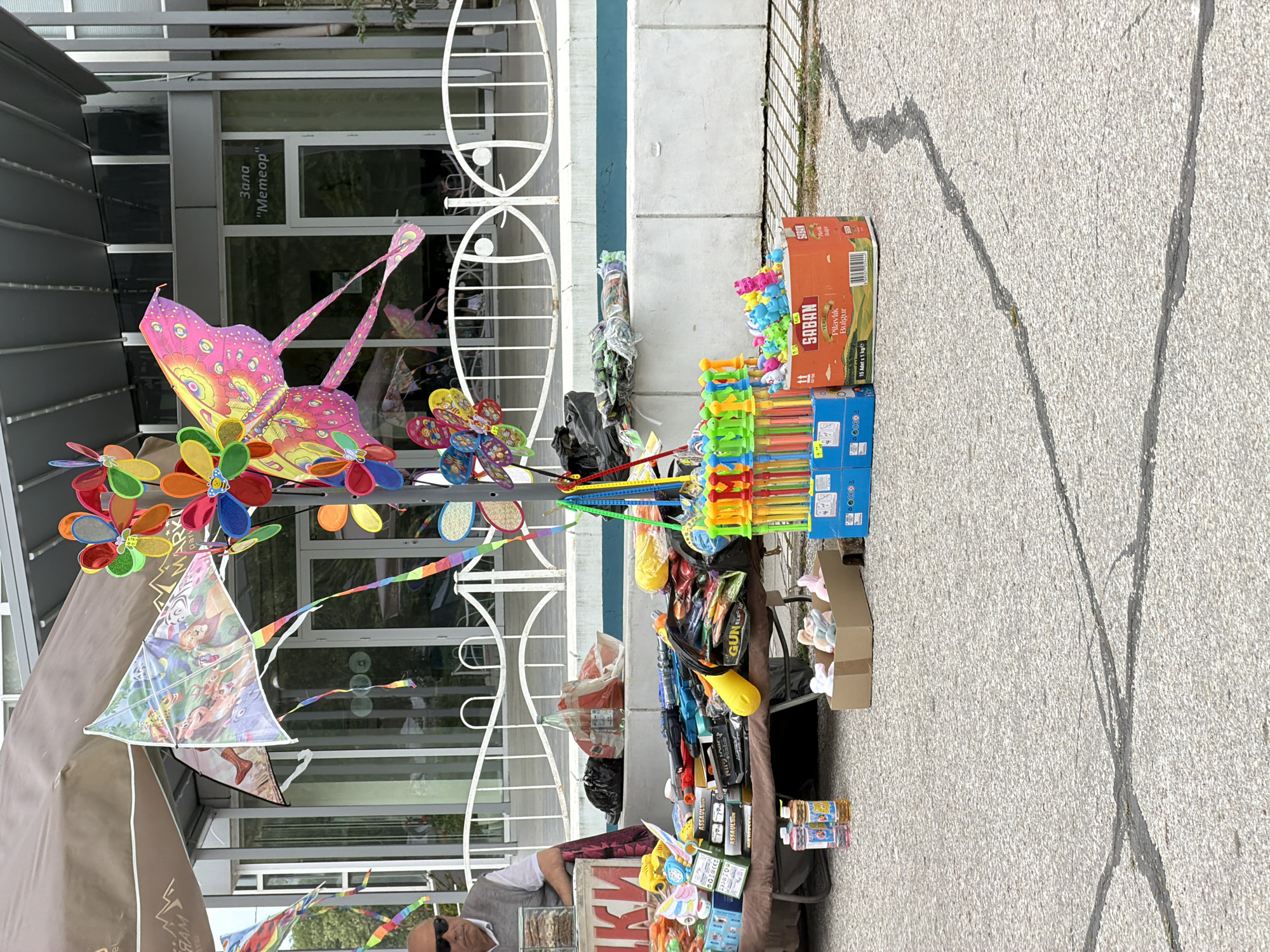 The image shows a street vendor's stall in Plovdiv, Bulgaria, filled with a variety of children's toys. Colorful kites, pinwheels, and plastic water guns are displayed on a table next to a building with a sign that reads "Зала 'Метеор'". The vendor's goods extend onto the pavement, including boxes of bulgur. The street surface shows cracks, indicating possible repairs.