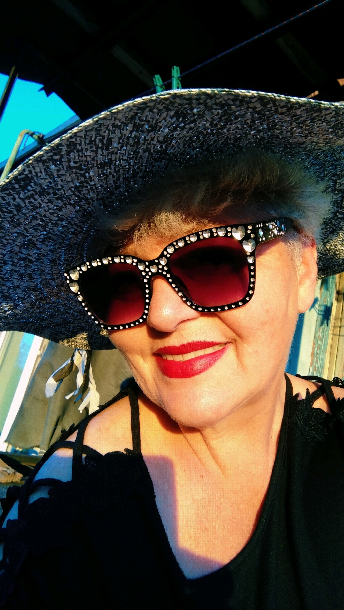 The image is a close-up, outdoor selfie of an elderly Caucasian woman wearing a wide-brimmed hat and large, rhinestone-encrusted sunglasses. She has short, greyish-blonde hair and is wearing bright red lipstick. She is smiling, and her head is tilted slightly to her left. She is wearing a black top with decorative shoulder straps. The background is out of focus, but appears to be a covered outdoor area with some material hanging in the background. The lighting suggests it is either early morning or late afternoon. The image does not contain any information that could be used to identify its location in Horishni Plavni, Ukraine.