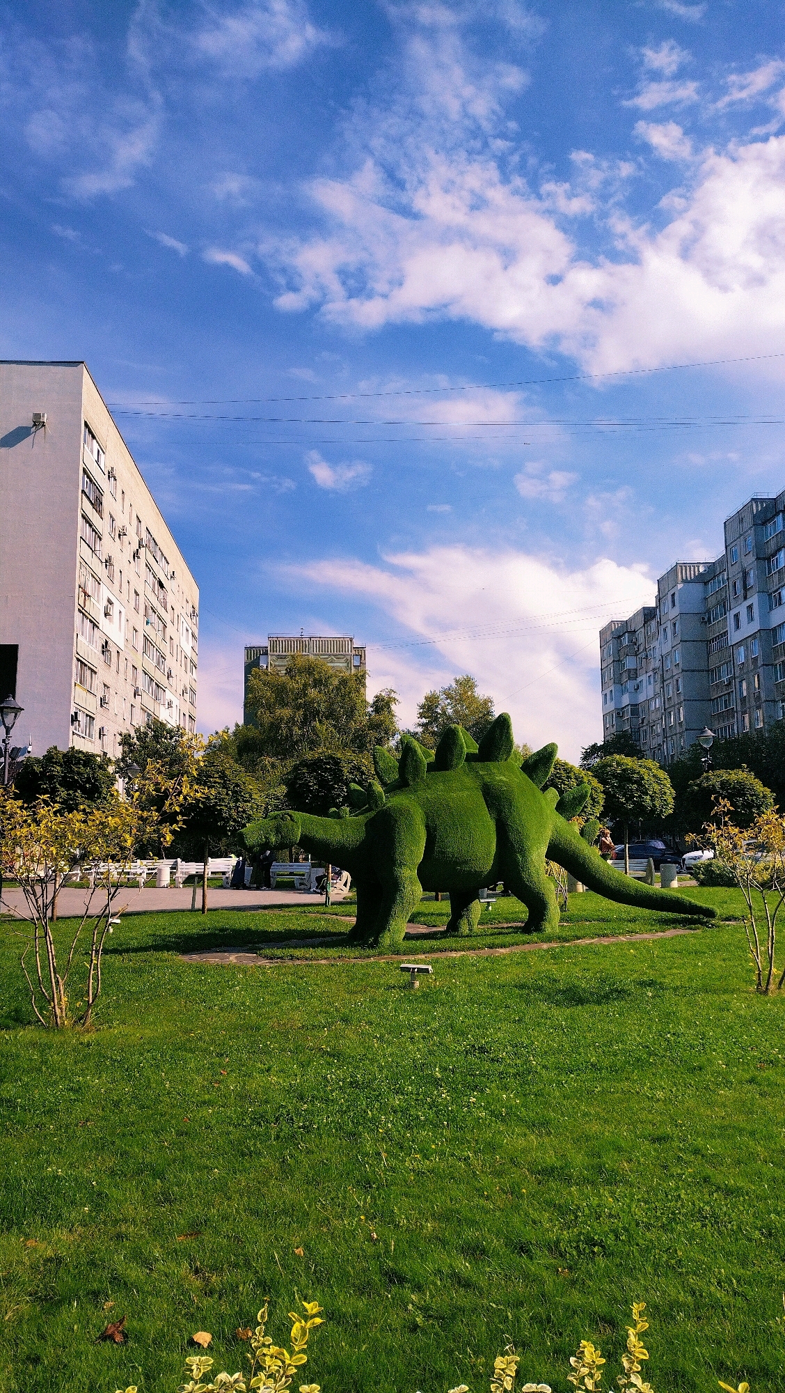 The image depicts a sunny day in the city of Horishni Plavni, Ukraine, with a vibrant blue sky dotted with white clouds. The foreground is dominated by a lush green lawn, interspersed with small yellow-leafed plants at the bottom. In the middle ground, a prominent topiary dinosaur, shaped like a Stegosaurus, stands as a whimsical centerpiece on the grass. Behind the dinosaur, several white park benches are visible, with a few people seated on them. To the left, a tall, multi-story apartment building stands, its facade a neutral color. Further in the background, more apartment buildings and trees create a cityscape. The overall scene suggests a public park or a well-maintained urban green space.