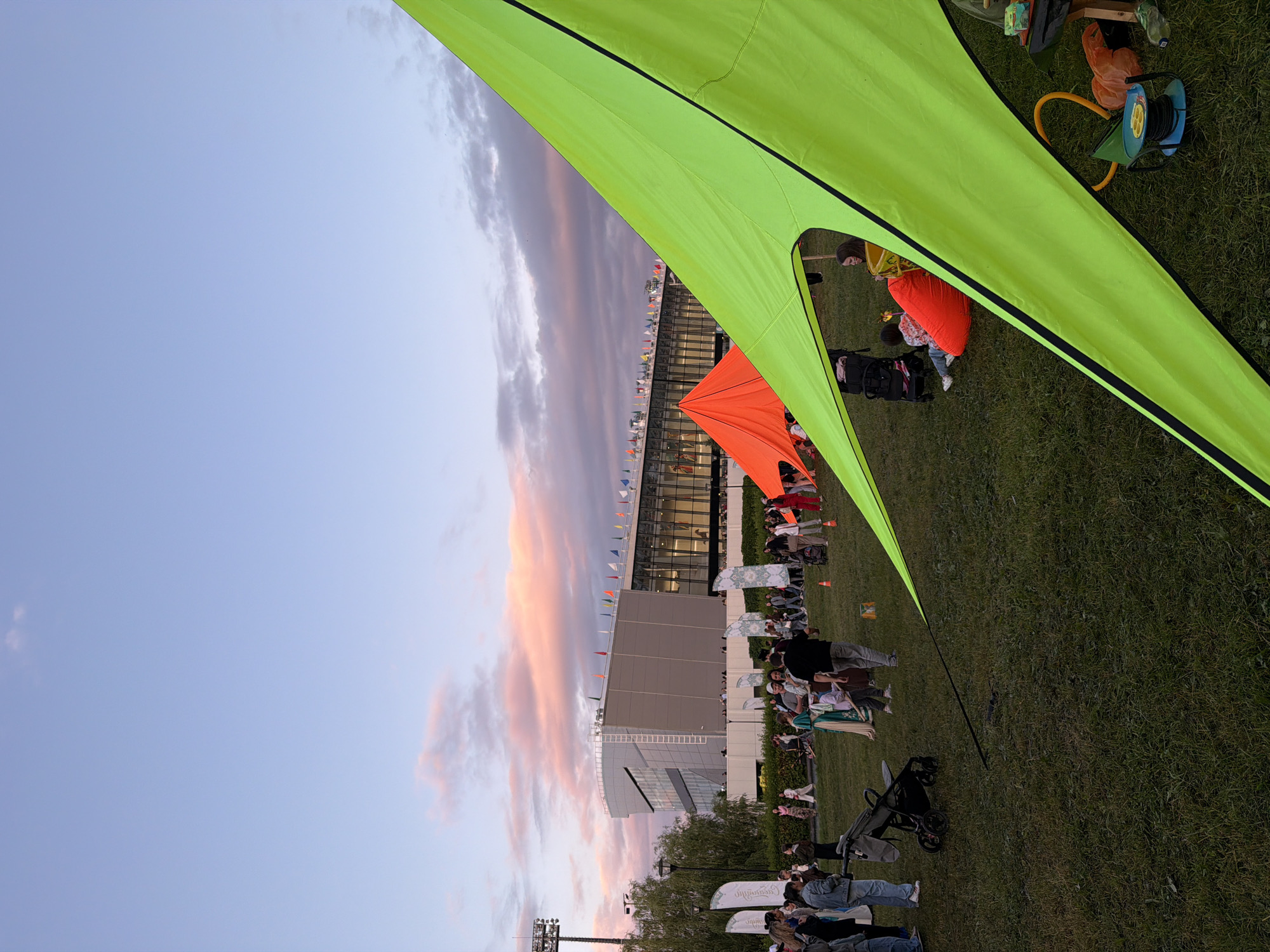 A wide shot captures an outdoor event at dusk, with a modern building and a stadium in the background. The sky is a gradient of light blue to pink and purple hues, indicating sunset. In the foreground, a large, bright lime-green tent dominates the right side of the frame, extending upwards and to the left. A smaller, bright orange tent is visible behind and to the left of the green tent.
The middle ground shows a grassy field with a gathering of people. Some individuals are walking, others are standing and talking, and a few are seated on the grass. Several white flags with geometric patterns are planted in the ground. A baby stroller is visible on the left side of the frame.
In the background, the modern building features large glass windows and a light-colored facade. A long row of small flags in various colors is mounted along the top of the building. A tall light tower stands to the far left. The overall scene suggests a festival, concert, or outdoor market taking place in a public space.