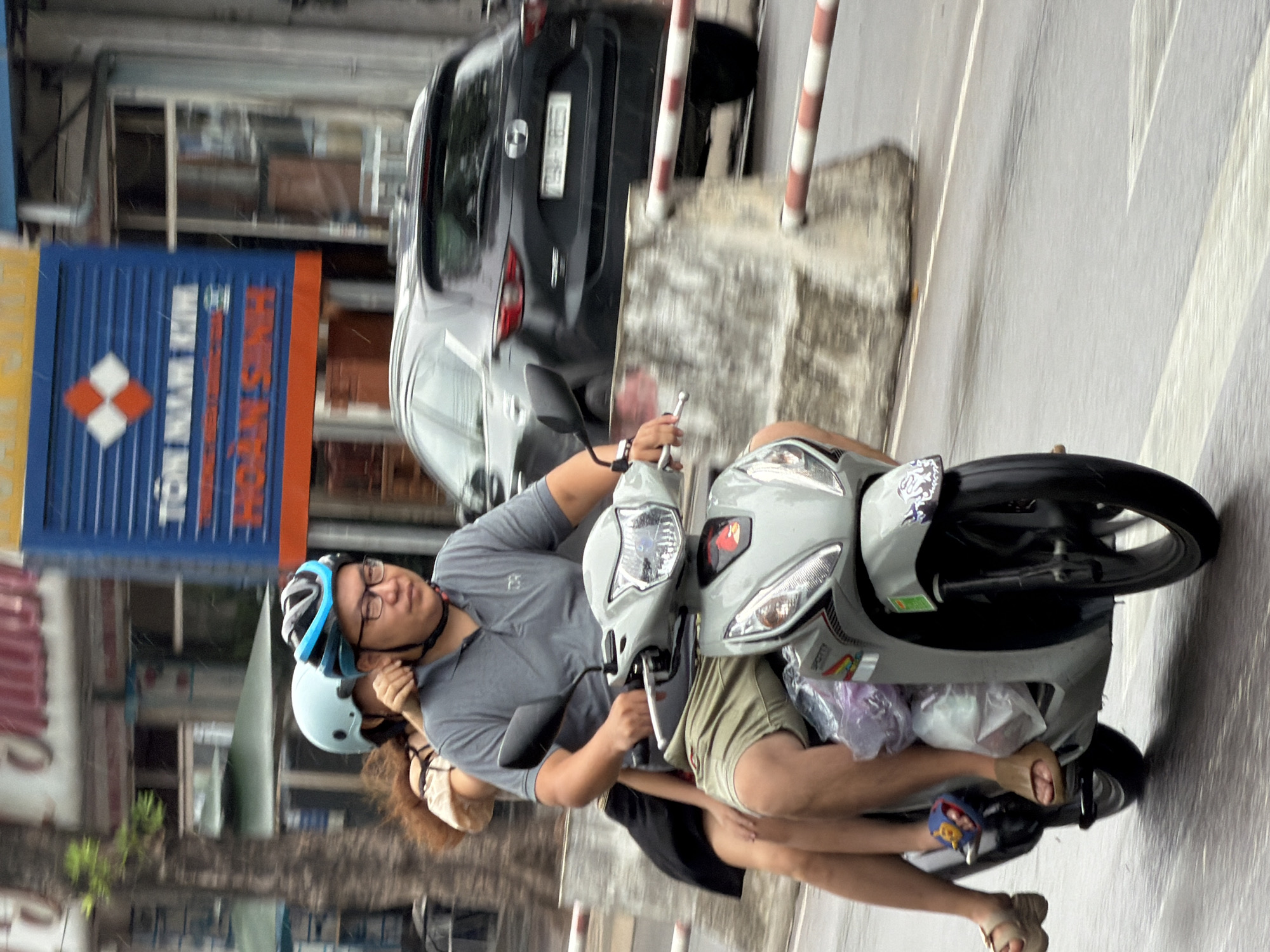 A medium shot captures a man and a woman riding on a scooter on a street in Nha Trang, Vietnam. The man, positioned in the front, wears a gray polo shirt, khaki shorts, and a blue and black helmet, and is looking straight ahead. He has glasses on his face. The woman, seated behind him, has long, dark hair and is wearing a beige top and a black skirt. She is wearing a light blue helmet and is holding onto the man. The scooter is gray and has a license plate visible. In the background, a dark-colored car is passing by. A building with a blue and yellow sign is also visible. The street appears wet, suggesting it may have recently rained. The overall scene depicts a typical street scene in a Vietnamese city.