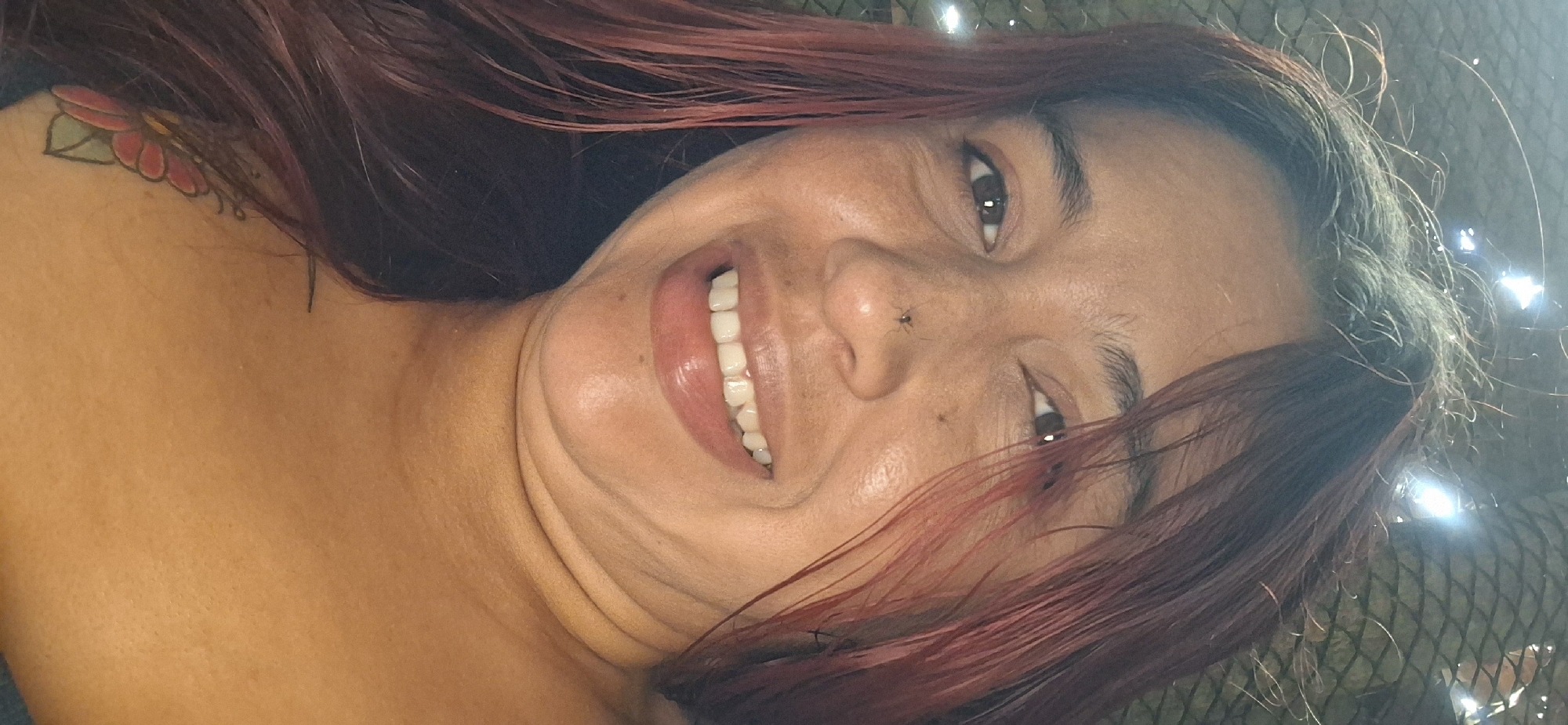 A close-up, rotated portrait captures a woman smiling broadly, conveying joy or amusement. Her dark hair, with visible reddish or purplish tints, frames her face and falls over her shoulders. She has dark brown eyes and a wide, open-mouthed smile, revealing a full set of white teeth. Distinct folds are visible around her chin and neck, accentuated by her expression and the angle of the shot. Notably, a small, dark insect, possibly a fly or spider, rests on the bridge of her nose. On her left shoulder/neck area, a colorful tattoo of a flower with red petals and green leaves is partially visible beneath her hair. Her skin has a warm, possibly tanned, complexion. The background is dark and appears to be a mesh or net-like structure, with several bright, reflective light spots suggesting strong illumination, potentially from sunlight or artificial sources. The setting is casual, consistent with a personal photograph taken in a brightly lit environment in Vallecito, Venezuela. No text is visible in the media.