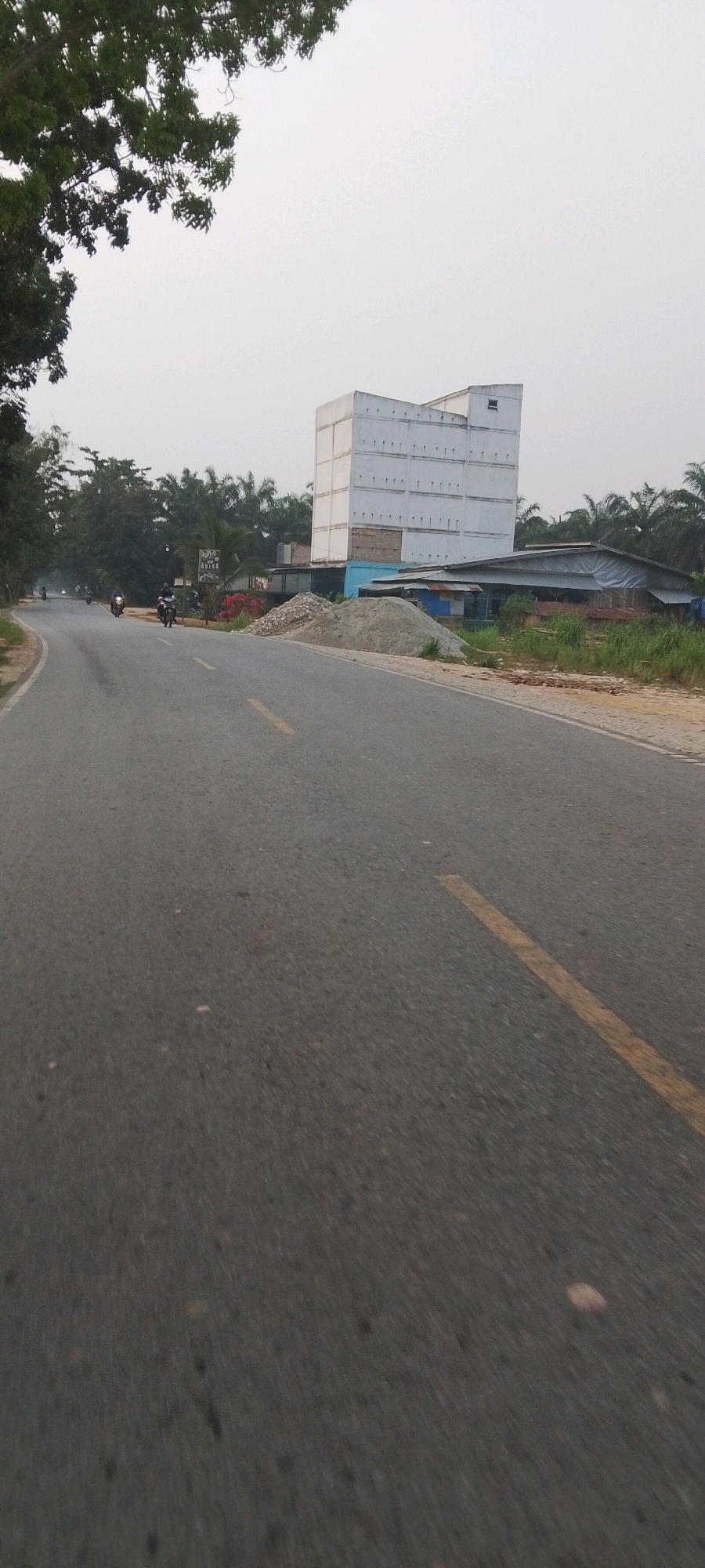 The image depicts a street scene in Lenggadai Hulu, Indonesia. A two-lane asphalt road, marked with a yellow line, curves to the left. Several motorcycles are traveling on the road, with riders wearing helmets. On the right side of the road, a multi-story white building with numerous windows dominates the background, situated behind a low-rise structure with a corrugated metal roof. A large pile of sand and gravel is present in front of the buildings. The left side of the road is lined with trees, including a large tree in the foreground with dense foliage. The sky is overcast, and the overall lighting suggests it is either early morning or late afternoon.