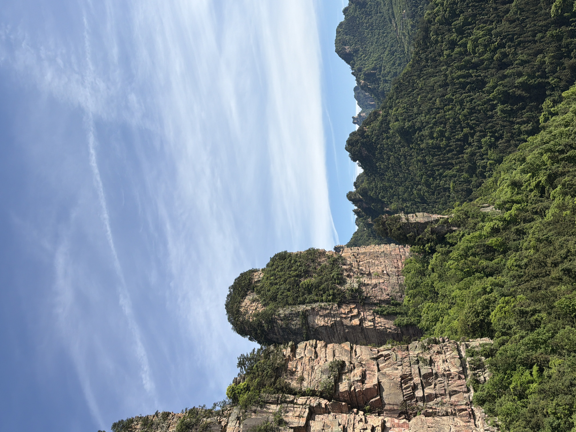 A low-angle, medium shot of a rocky mountain peak with a partially clear blue sky, likely taken in the area around Zhonghu, China. The mountain face is comprised of layered reddish-brown sedimentary rock formations, with a capping of dense green foliage, suggesting lush vegetation typical of the area's temperate climate. The layering of the rock is distinct, and the green foliage indicates a well-vegetated area. The sky is blue with thin, wispy cloud formations that run parallel to the vertical orientation of the image. The overall scene is characteristic of the landscape in the region.